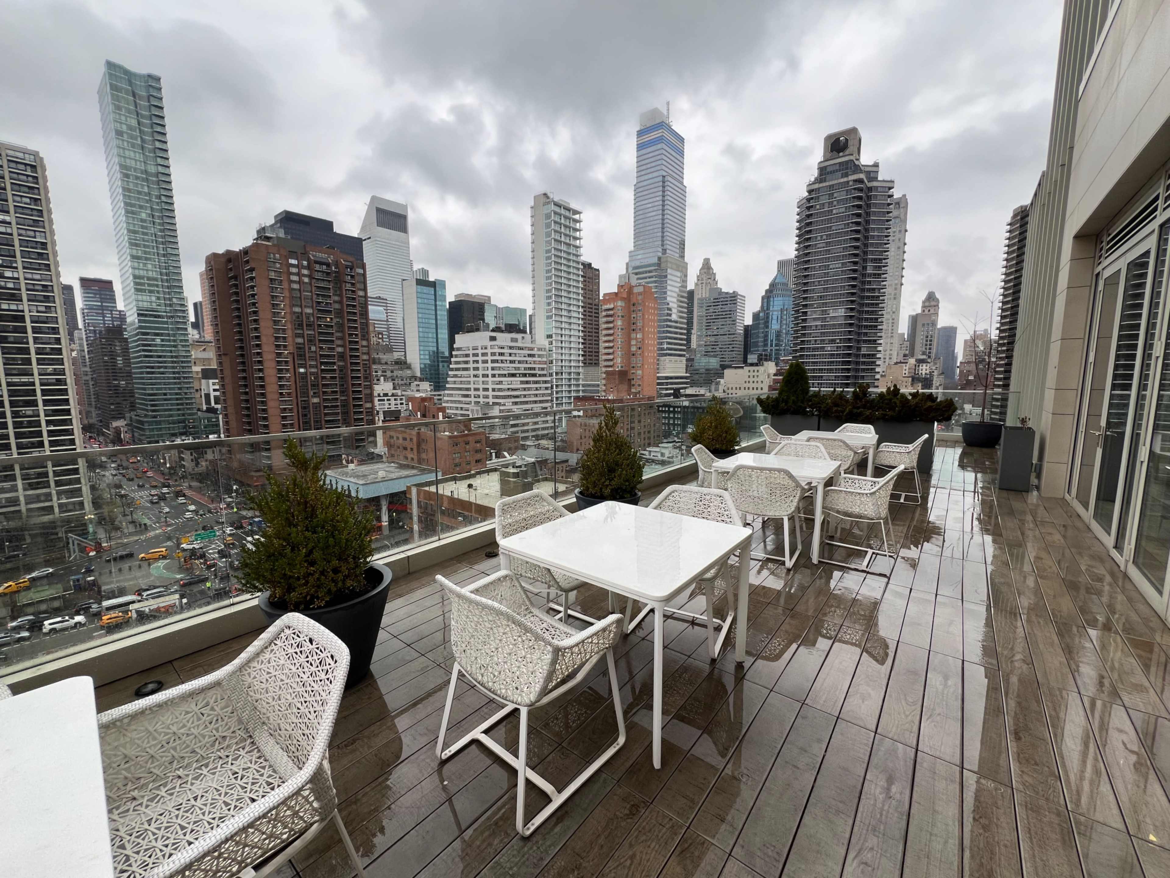 A rooftop terrace features white tables and chairs overlooking a city skyline with tall buildings under a cloudy sky.