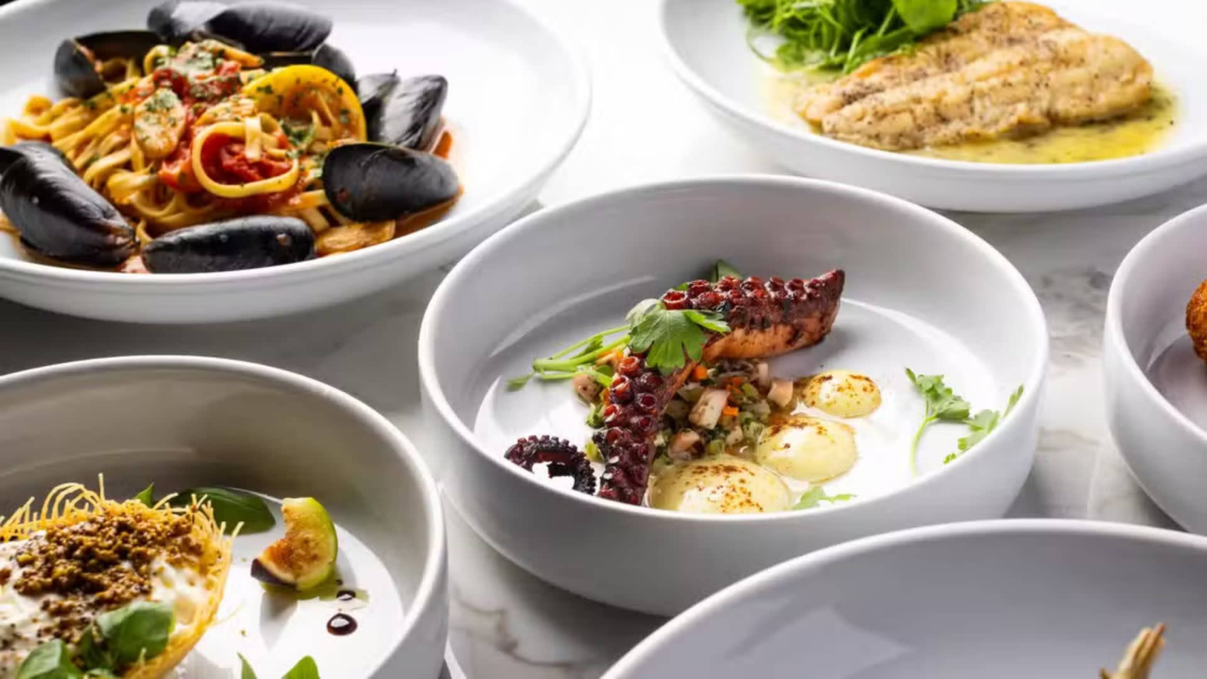 The image showcases a variety of dishes including pasta with seafood, a grilled fish fillet, and a plate featuring octopus with garnishes, all arranged on a marble surface.