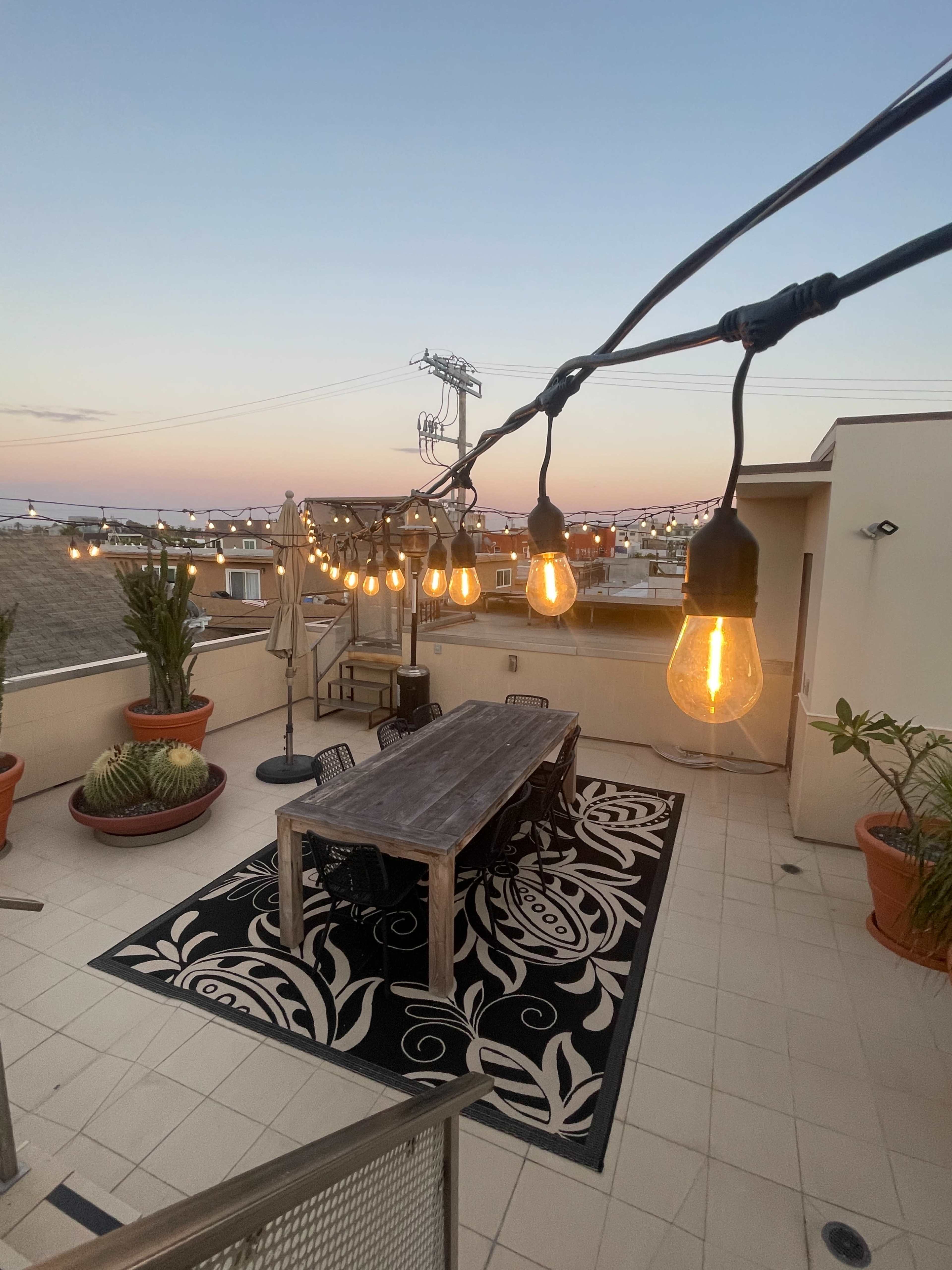 A rooftop patio features a wooden dining table surrounded by chairs, with string lights overhead and potted plants nearby, set against a twilight sky.