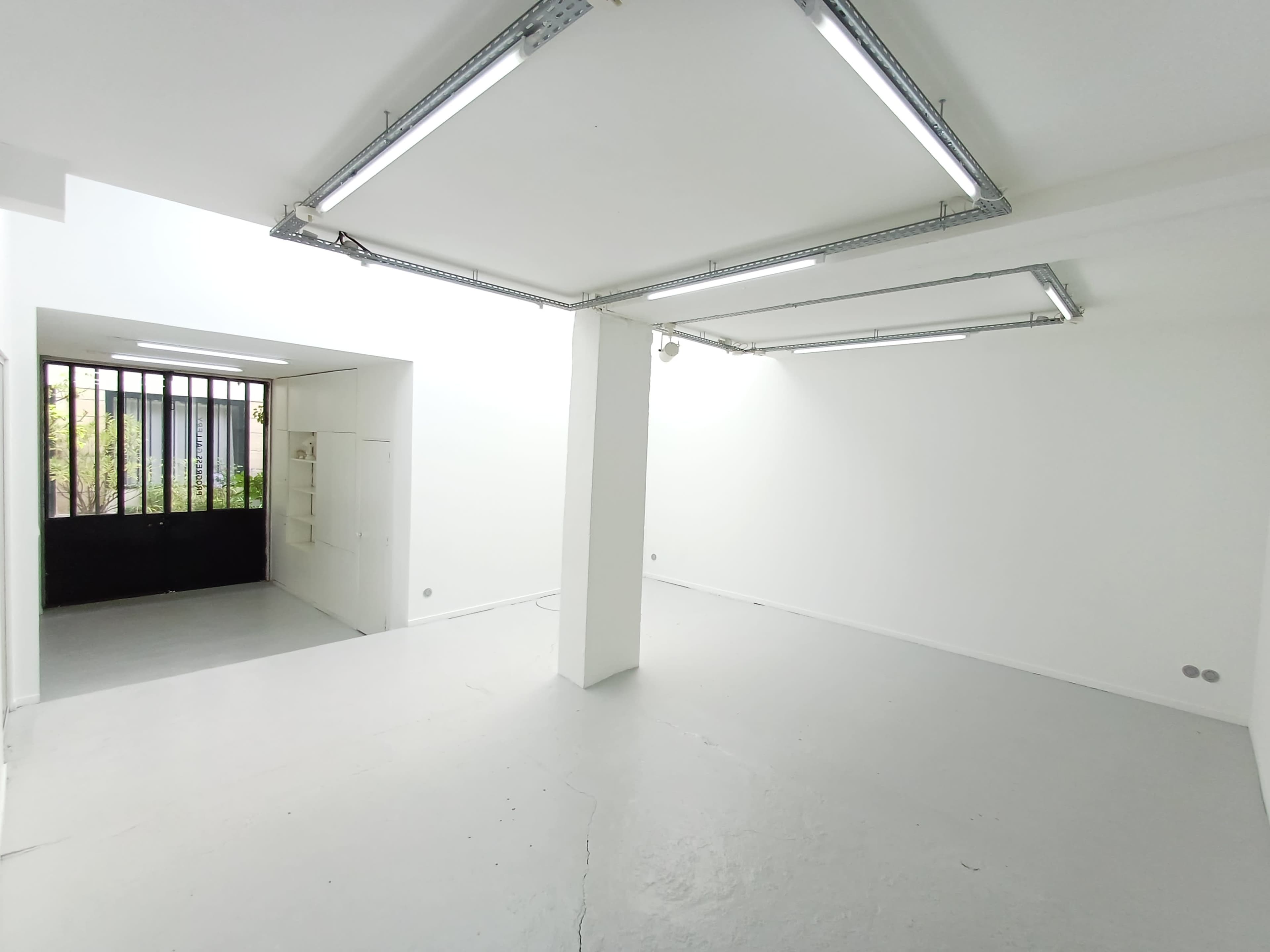The image shows a spacious, empty room with white walls and overhead lighting, leading to an exterior area with a barred window.