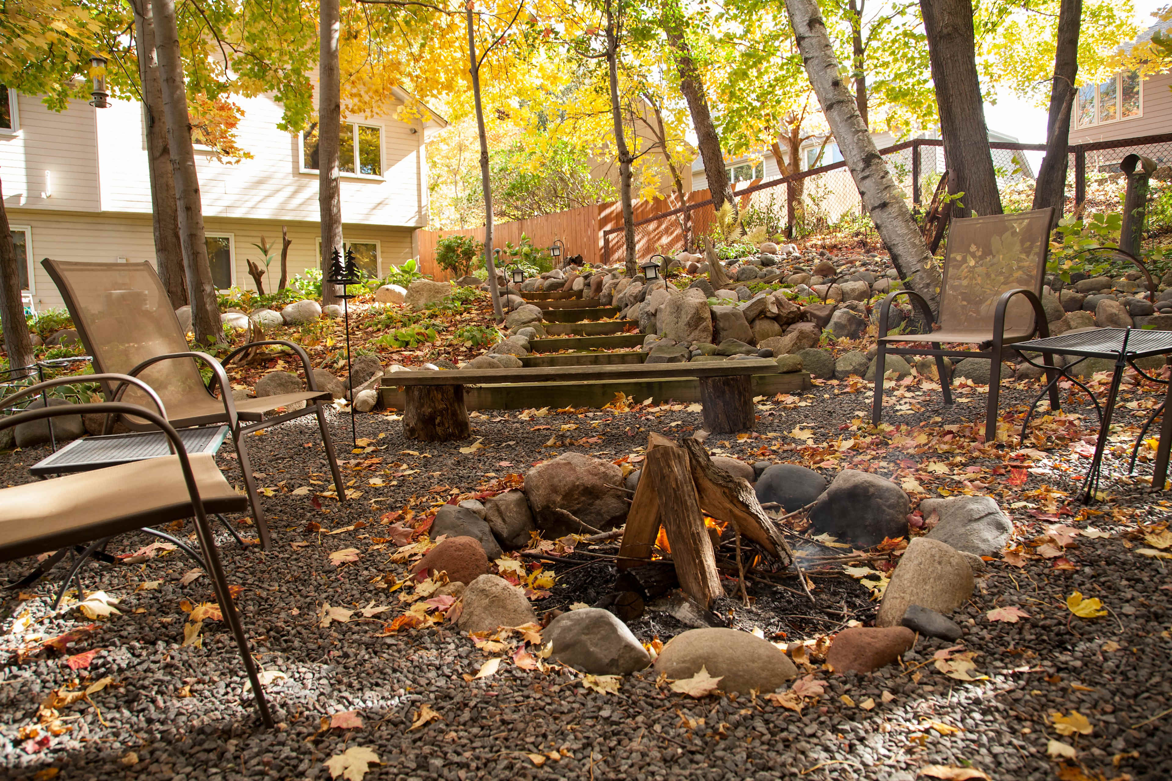 A circular fire pit surrounded by wooden logs and stones is set in a backyard garden adorned with autumn leaves and stone pathways.