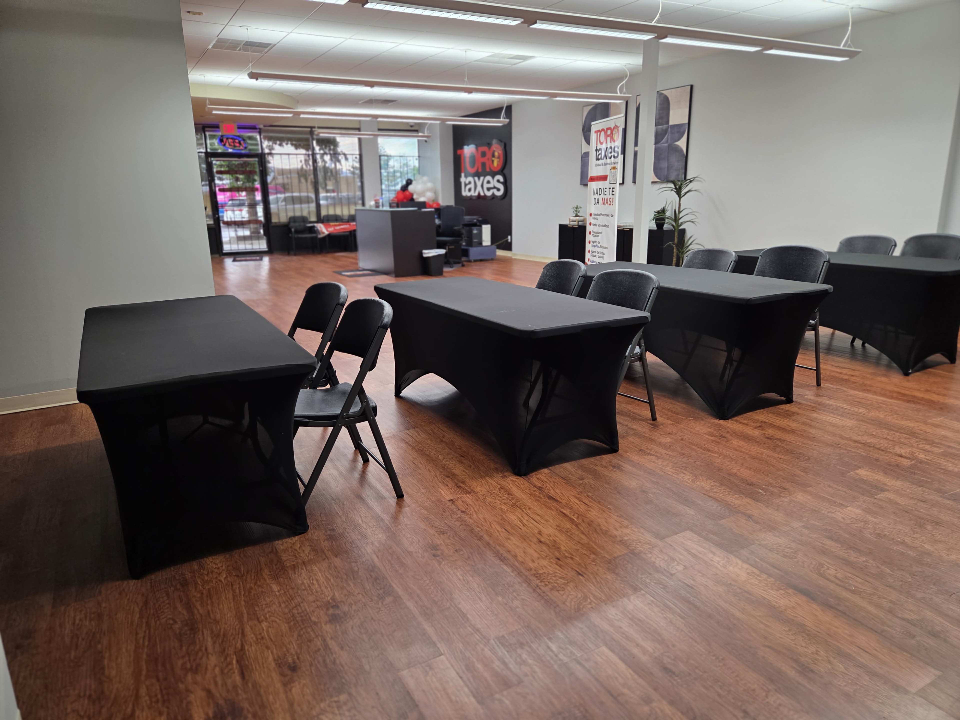 The image shows a room with multiple black tables and chairs arranged neatly, with a reception area visible in the background.