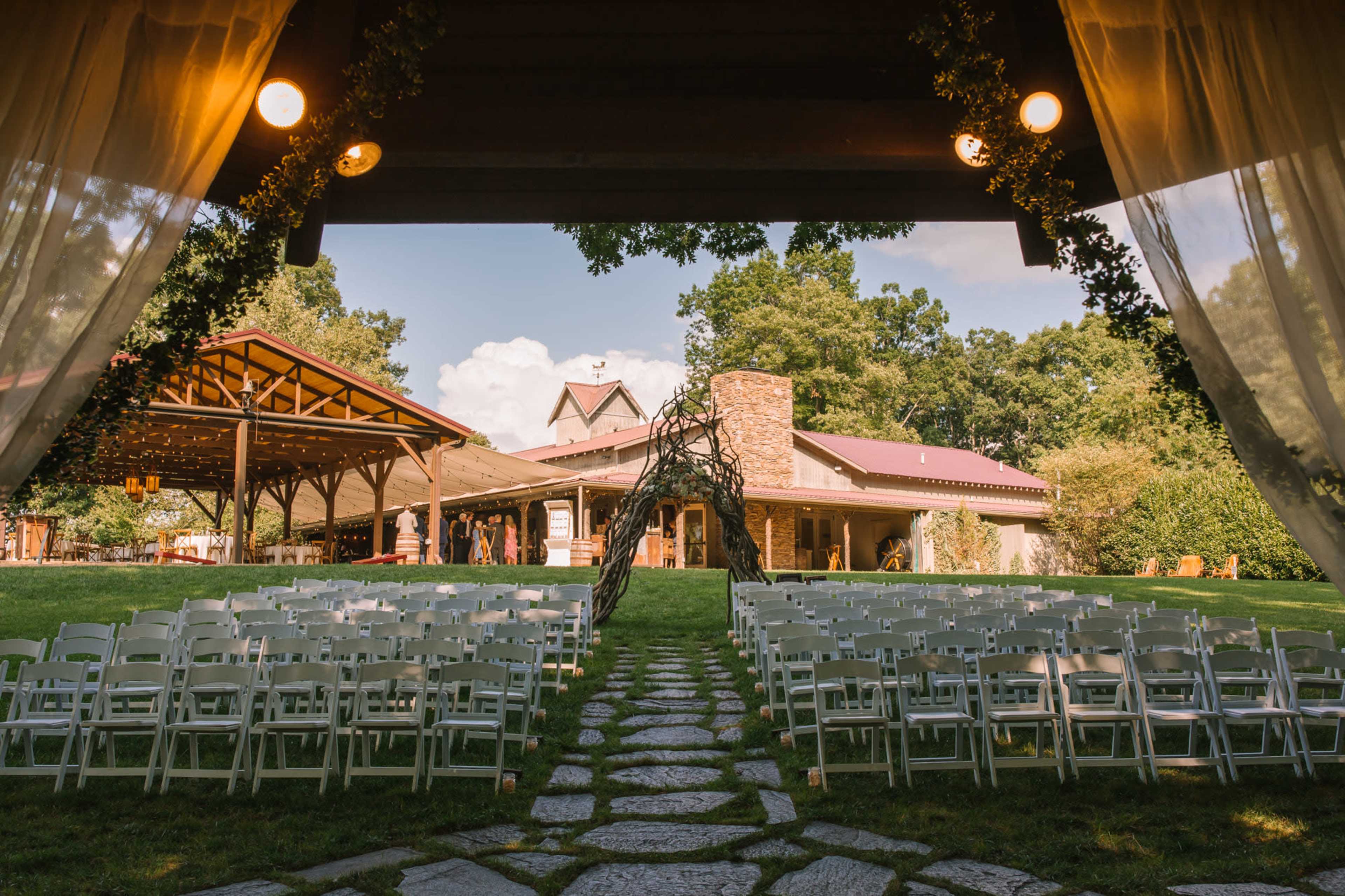 A pathway of stone leads to an outdoor event space surrounded by chairs set up for a gathering in front of a building with a sloped roof and large windows.
