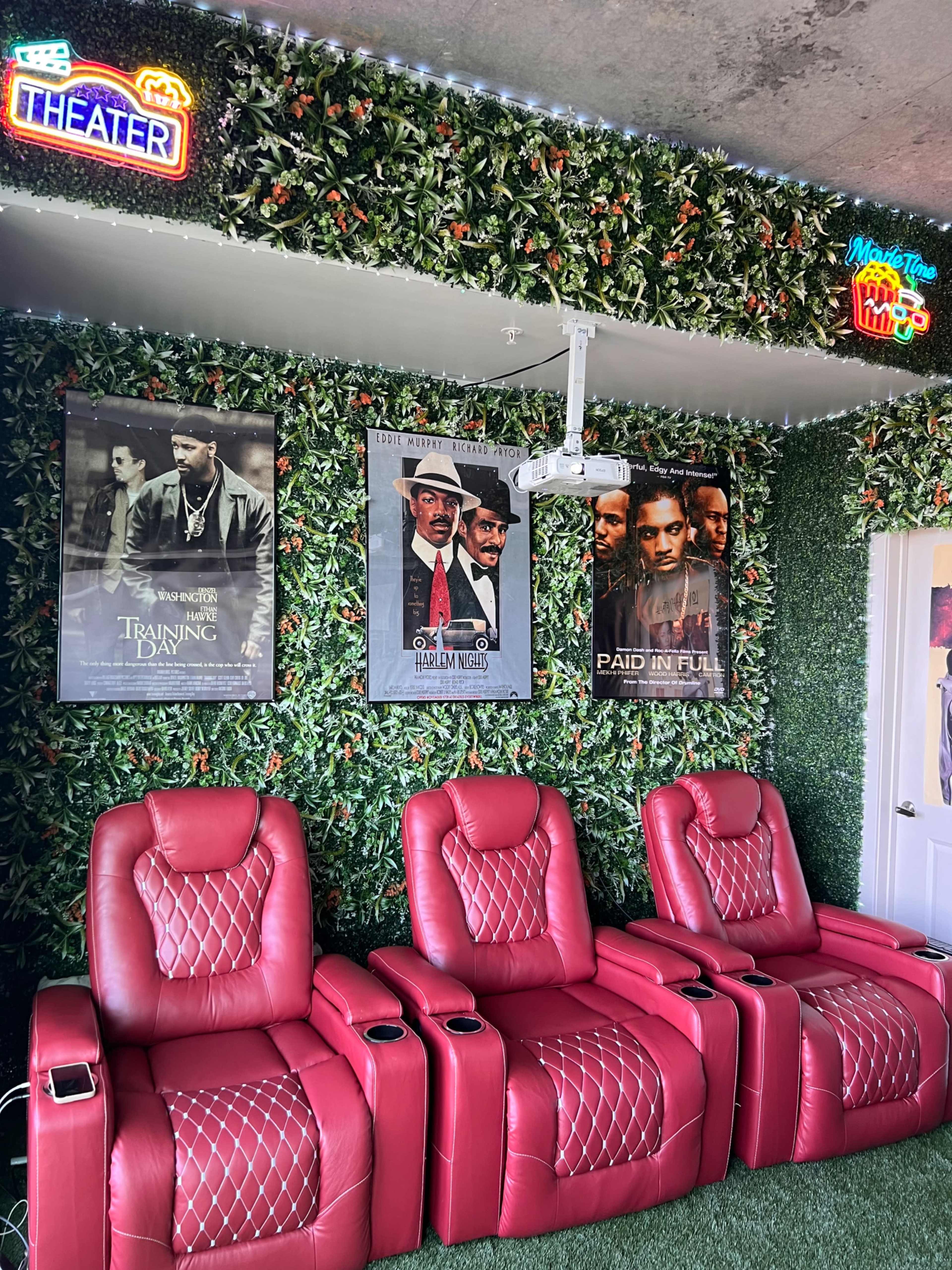The image shows a home theater area with red reclining chairs, posters of movies on a leafy green wall, and neon signage.
