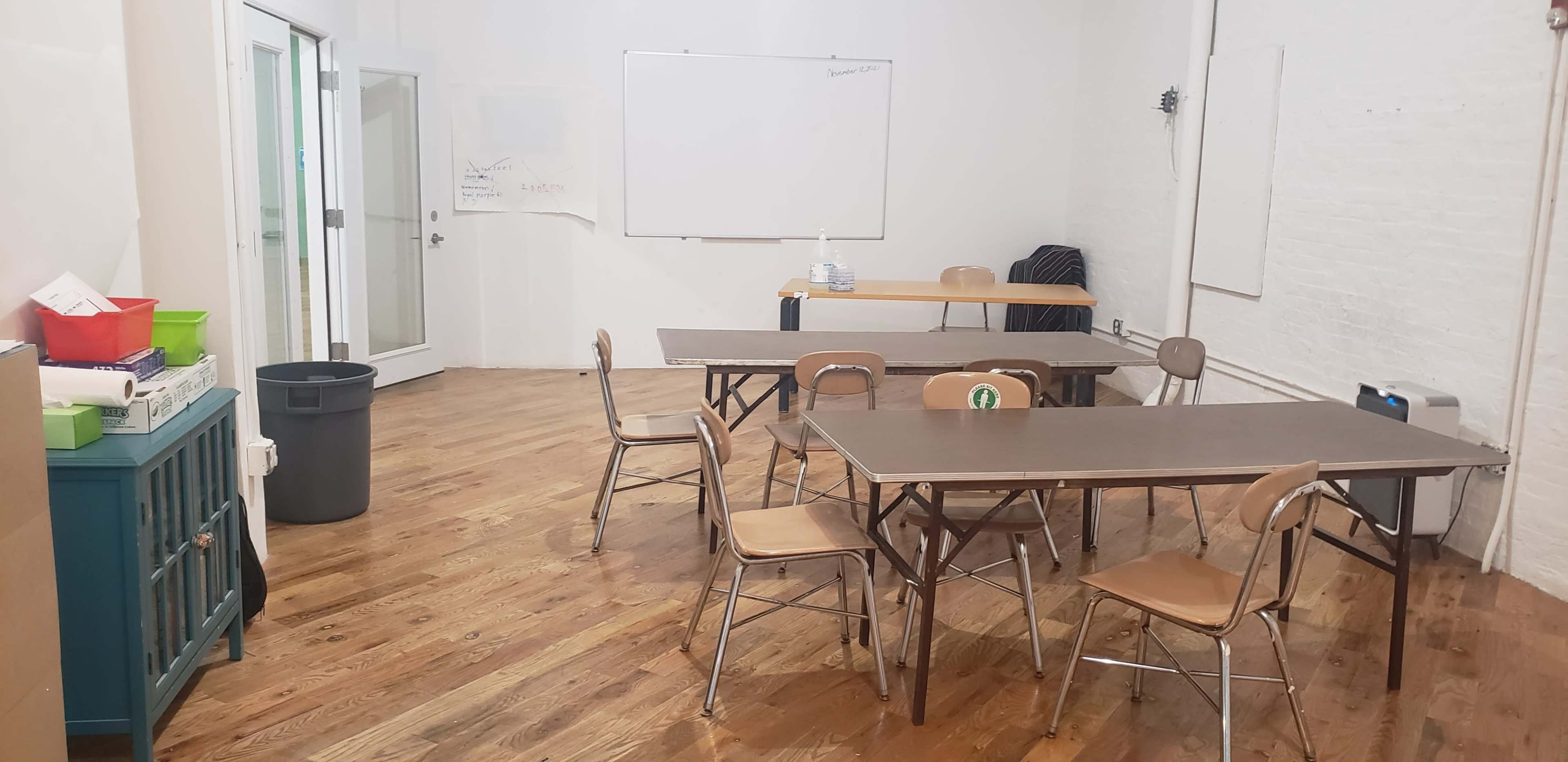 The image shows a classroom with several wooden tables and chairs, a trash bin, and a whiteboard on the wall in a bright, spacious setting.