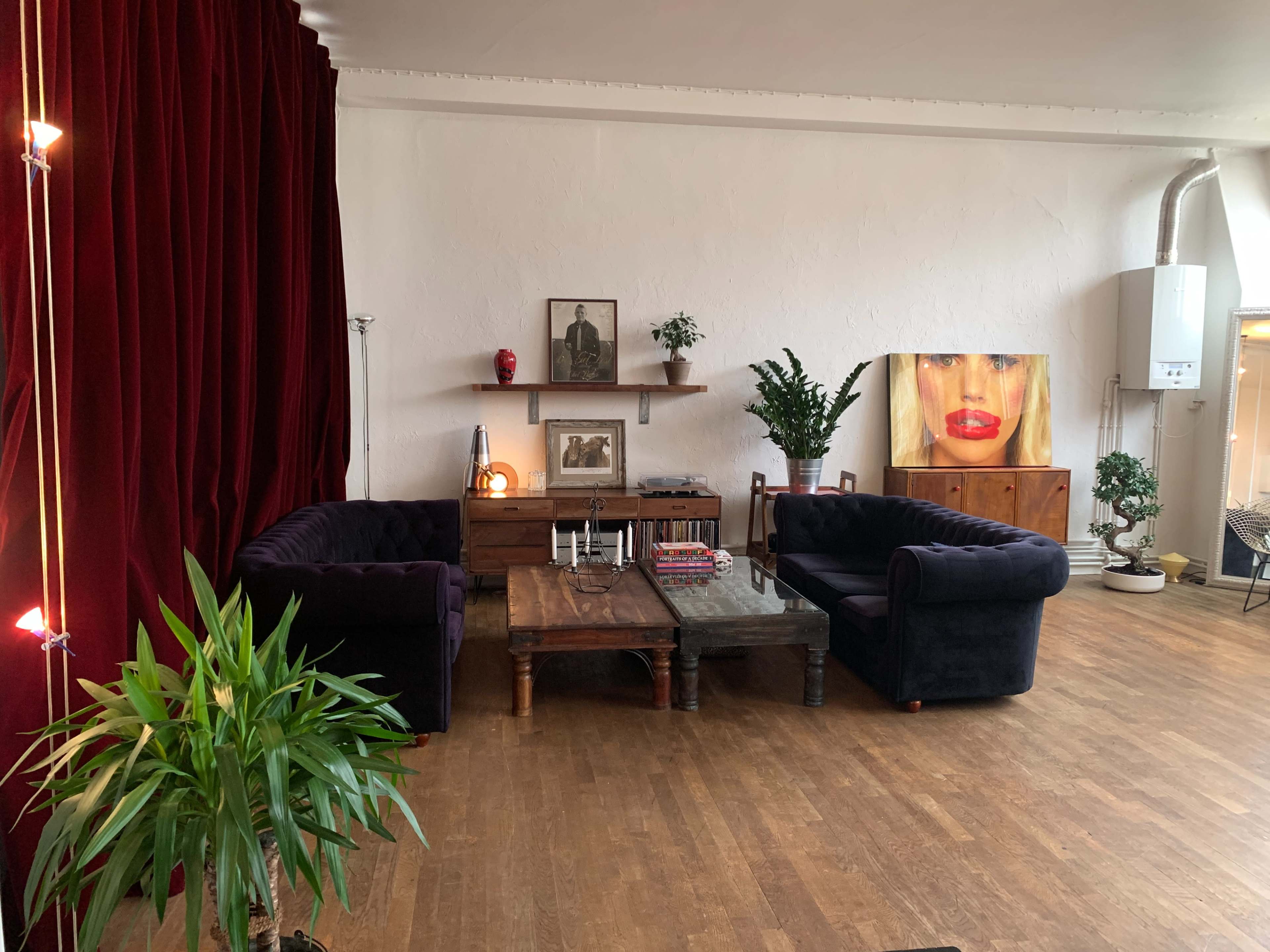 A cozy living space with two dark couches facing a wooden coffee table, surrounded by plants and a red curtain backdrop, featuring artwork on the walls.