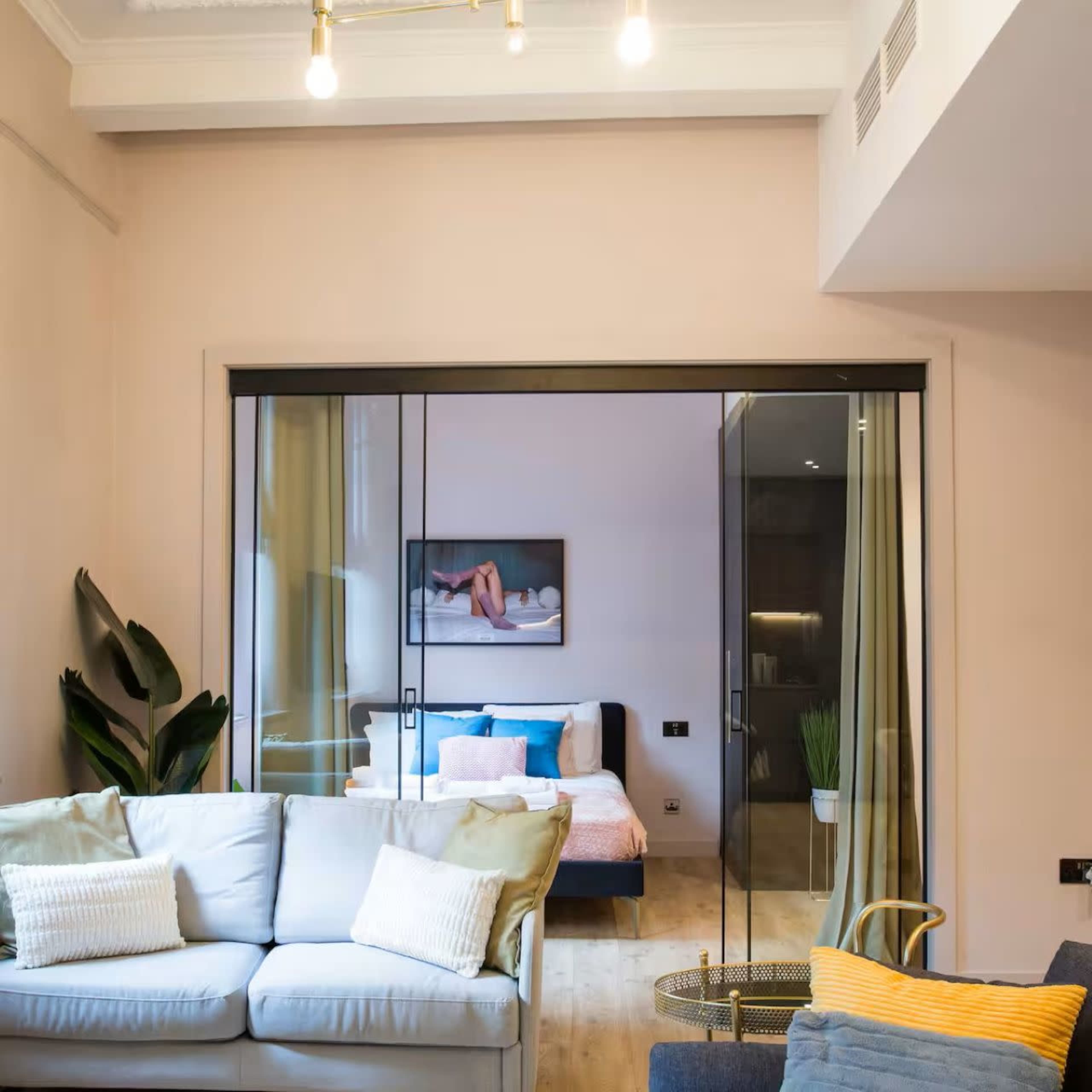 The image shows a modern living space featuring a soft sofa in the foreground and a glass partition leading to a bedroom with a blue and pink bedspread.