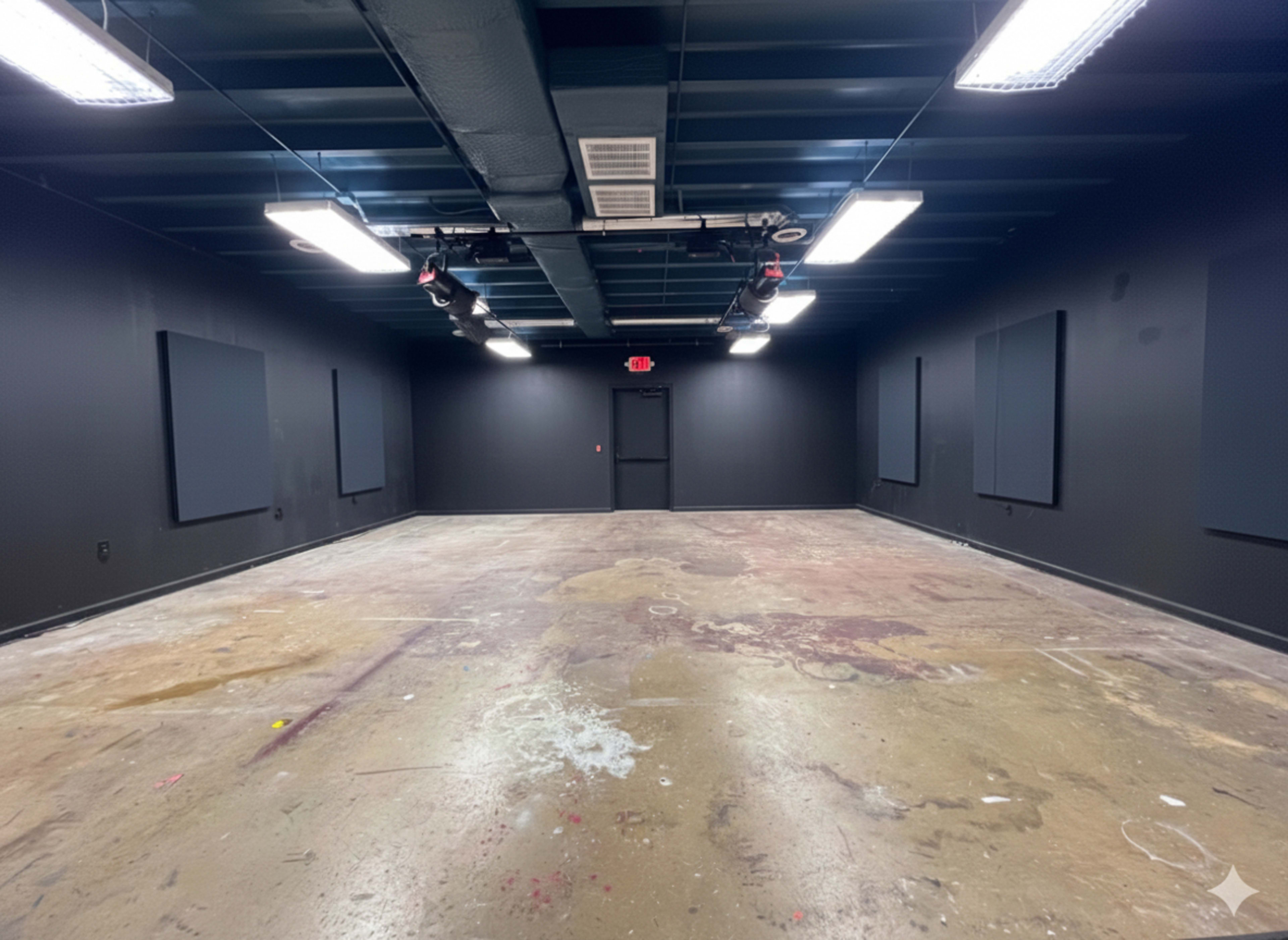 The image shows a large, empty room with dark walls, an unpainted concrete floor, and soundproof panels on the walls.