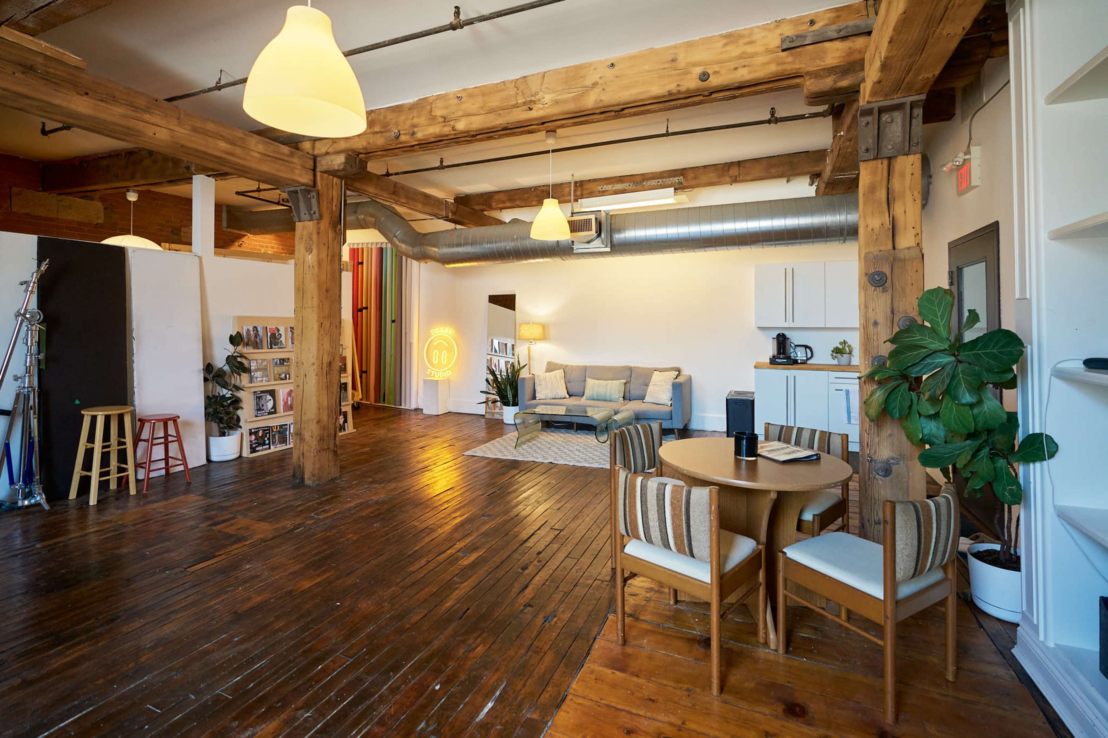 The image shows a spacious, open-concept studio with wooden beams, a cozy seating area, and a dining table set up near a kitchenette.