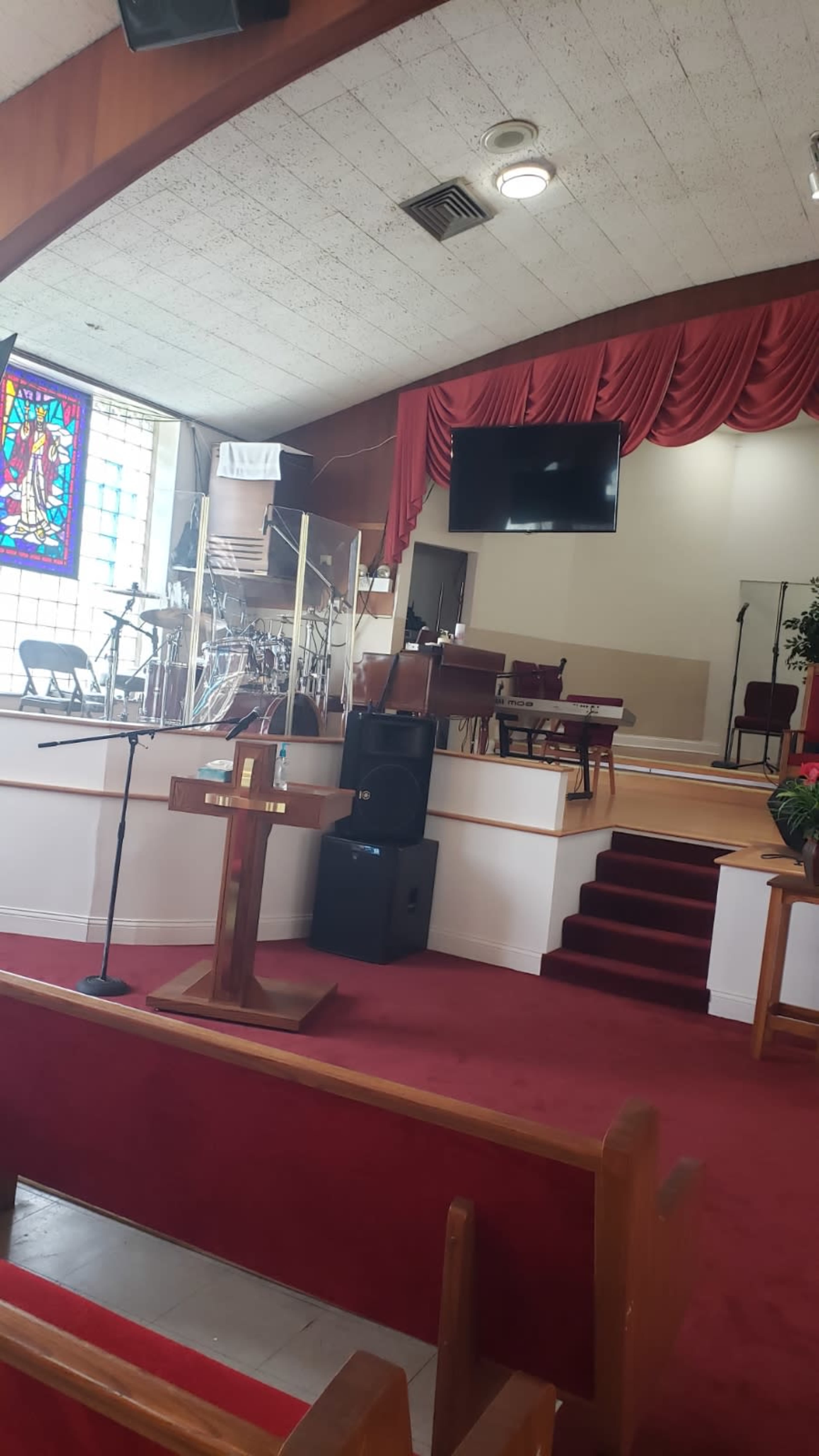 The image shows the interior of a church with a wooden pulpit, stained glass window, and a stage area equipped with musical instruments and a large screen.