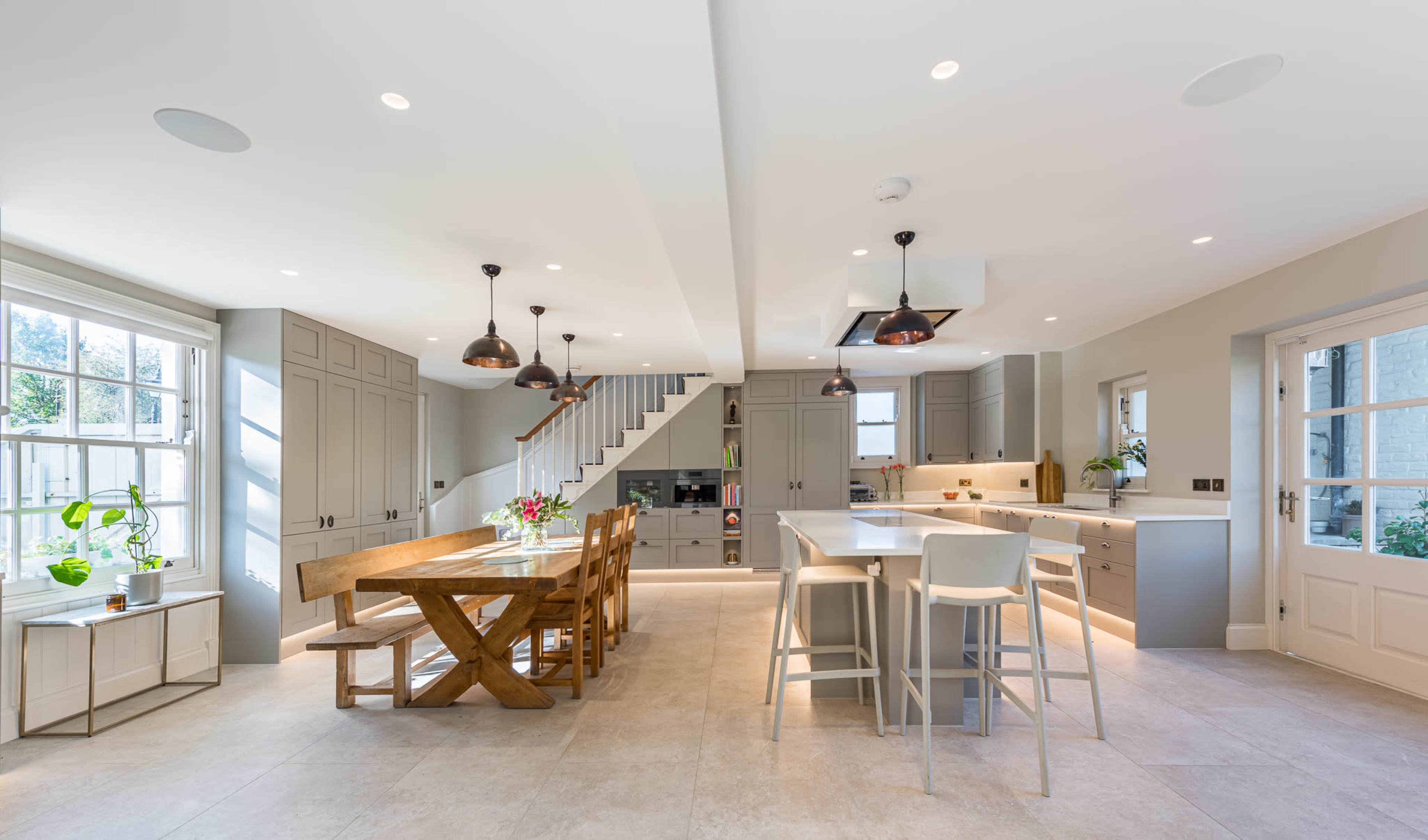 The image shows a modern kitchen and dining area featuring a large wooden dining table, white bar stools, and a staircase leading to an upper level.