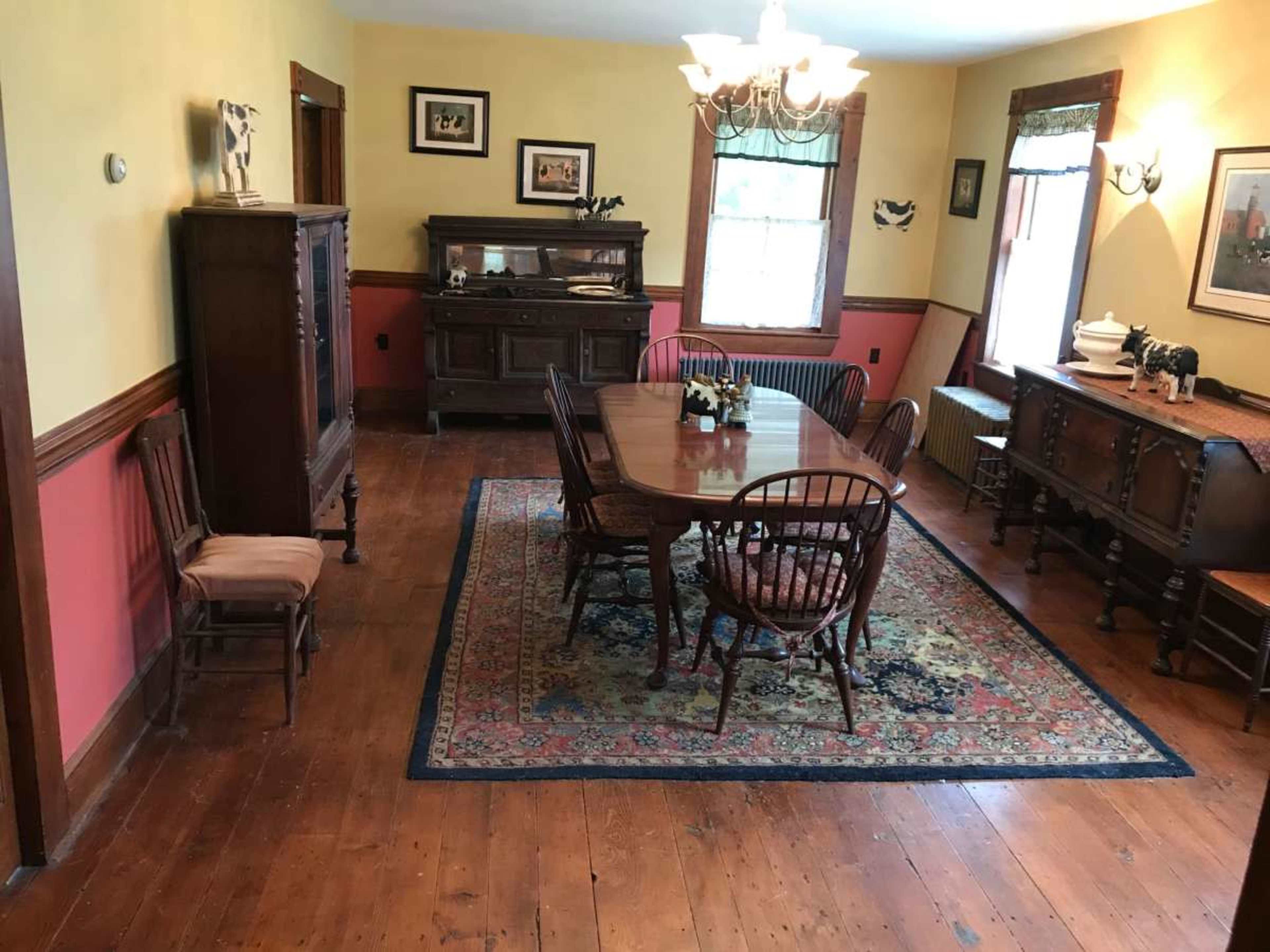 The image shows a dining room with a table set for several people, surrounded by wooden chairs, a sideboard, and vintage decor against yellow and red walls.