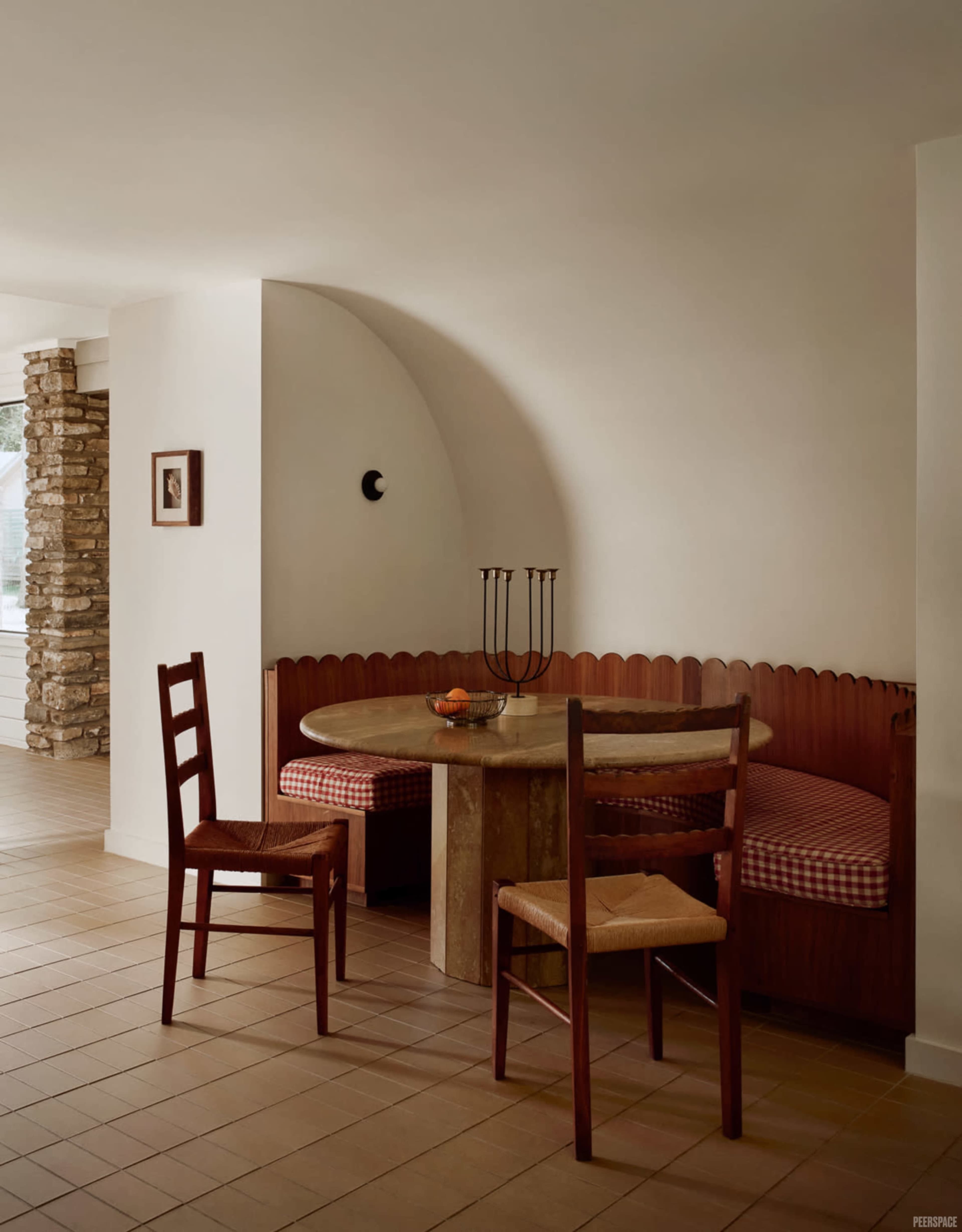 A nook featuring a circular marble table with a scalloped wooden bench and two wooden chairs is set against a plain wall.