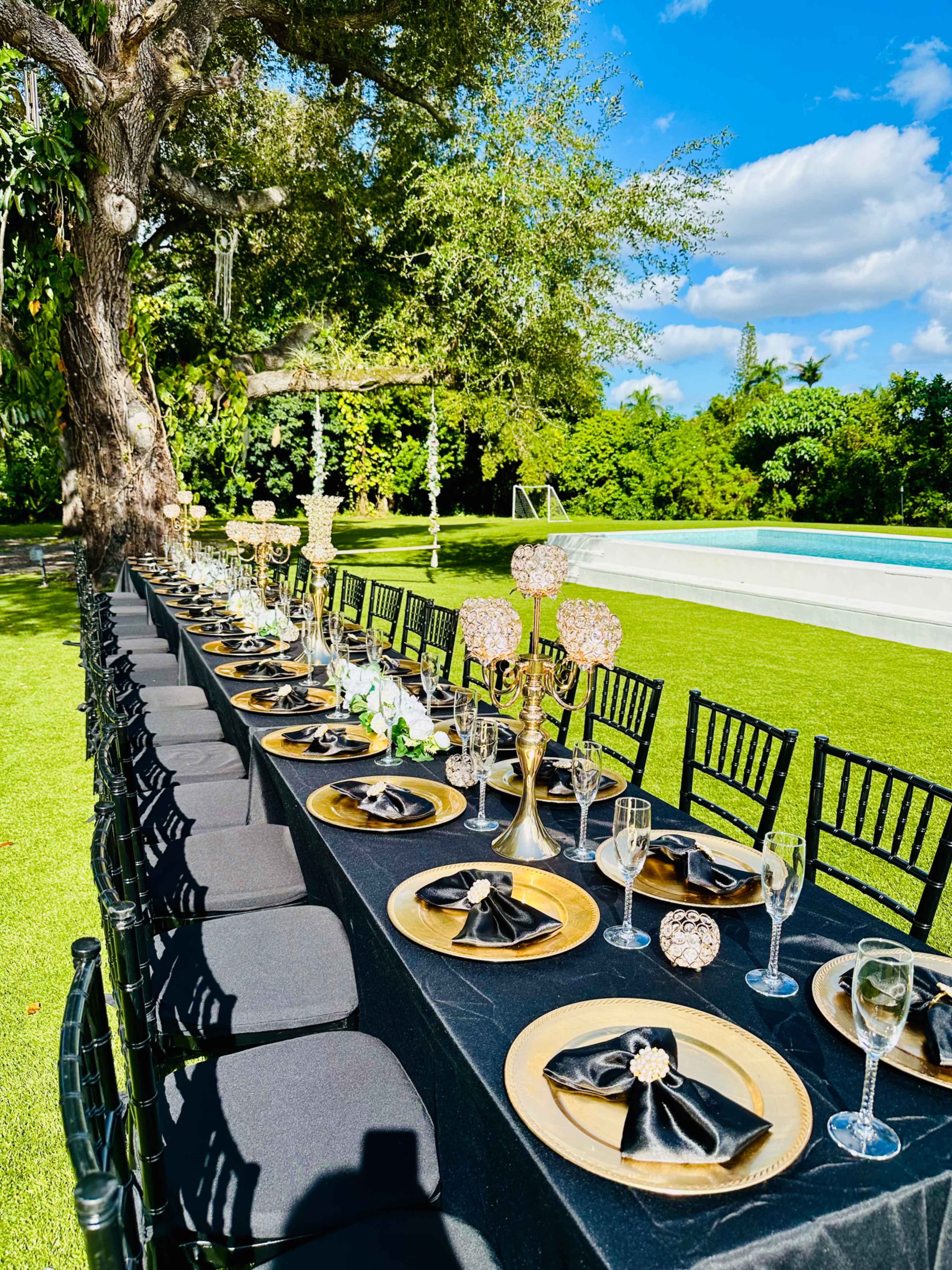 A long outdoor dining table is elegantly set with black and gold tableware under a clear blue sky, surrounded by lush greenery.