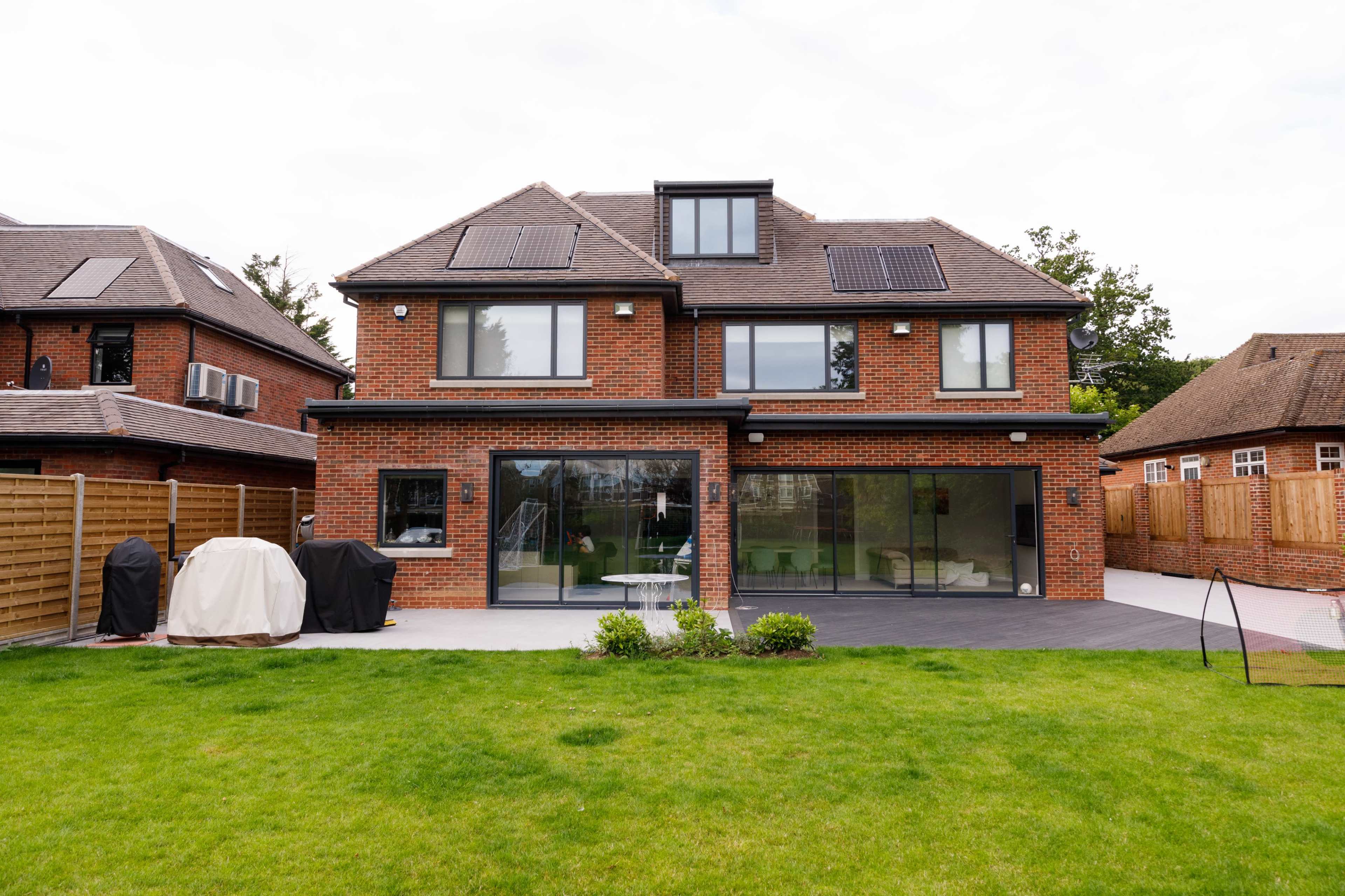 A modern two-story brick house with large glass windows and a landscaped backyard featuring a grassy lawn, patio, and outdoor furniture.