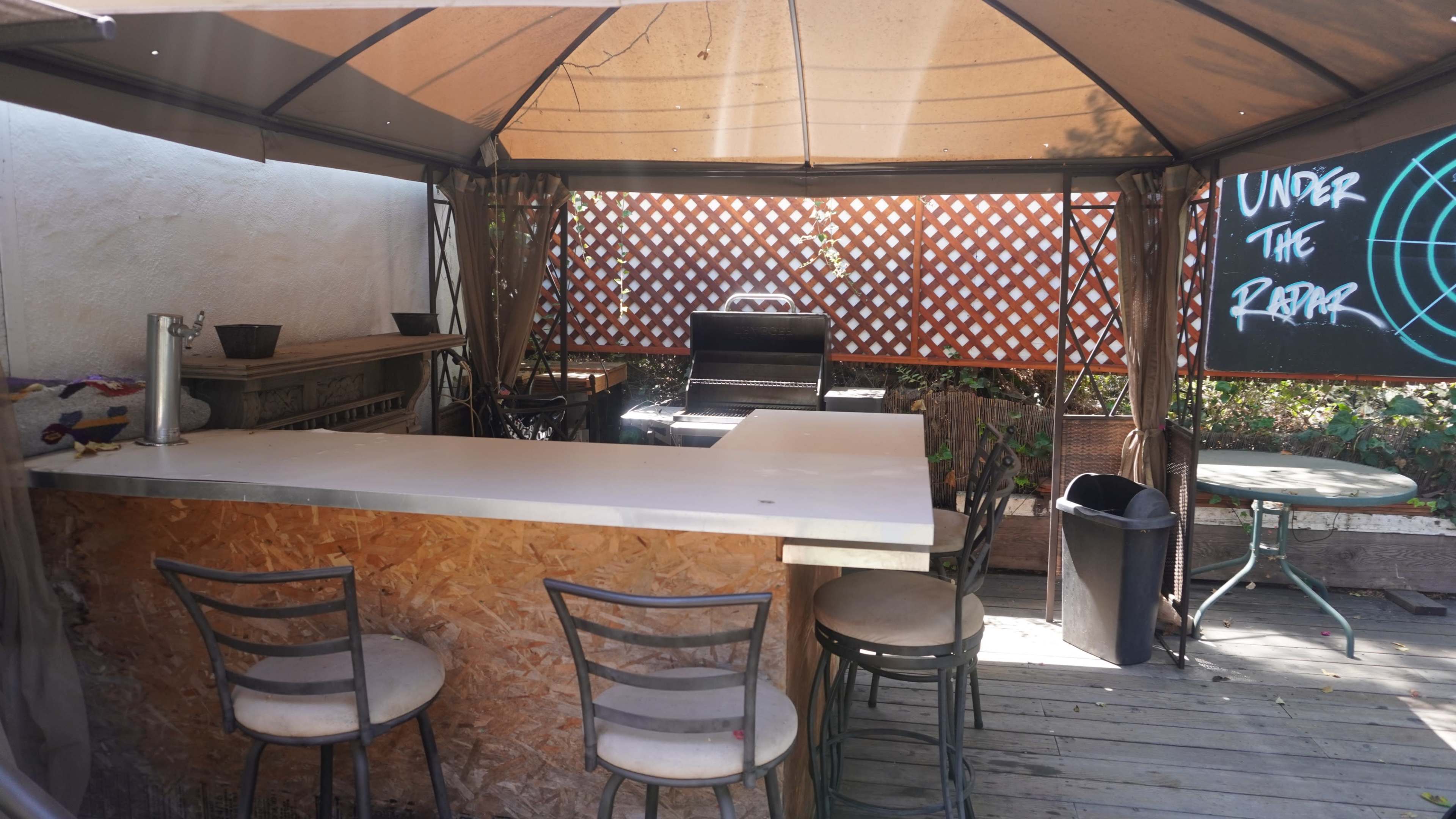 An outdoor bar area with a covered gazebo, featuring a countertop, several stools, and a barbecue grill in the background.