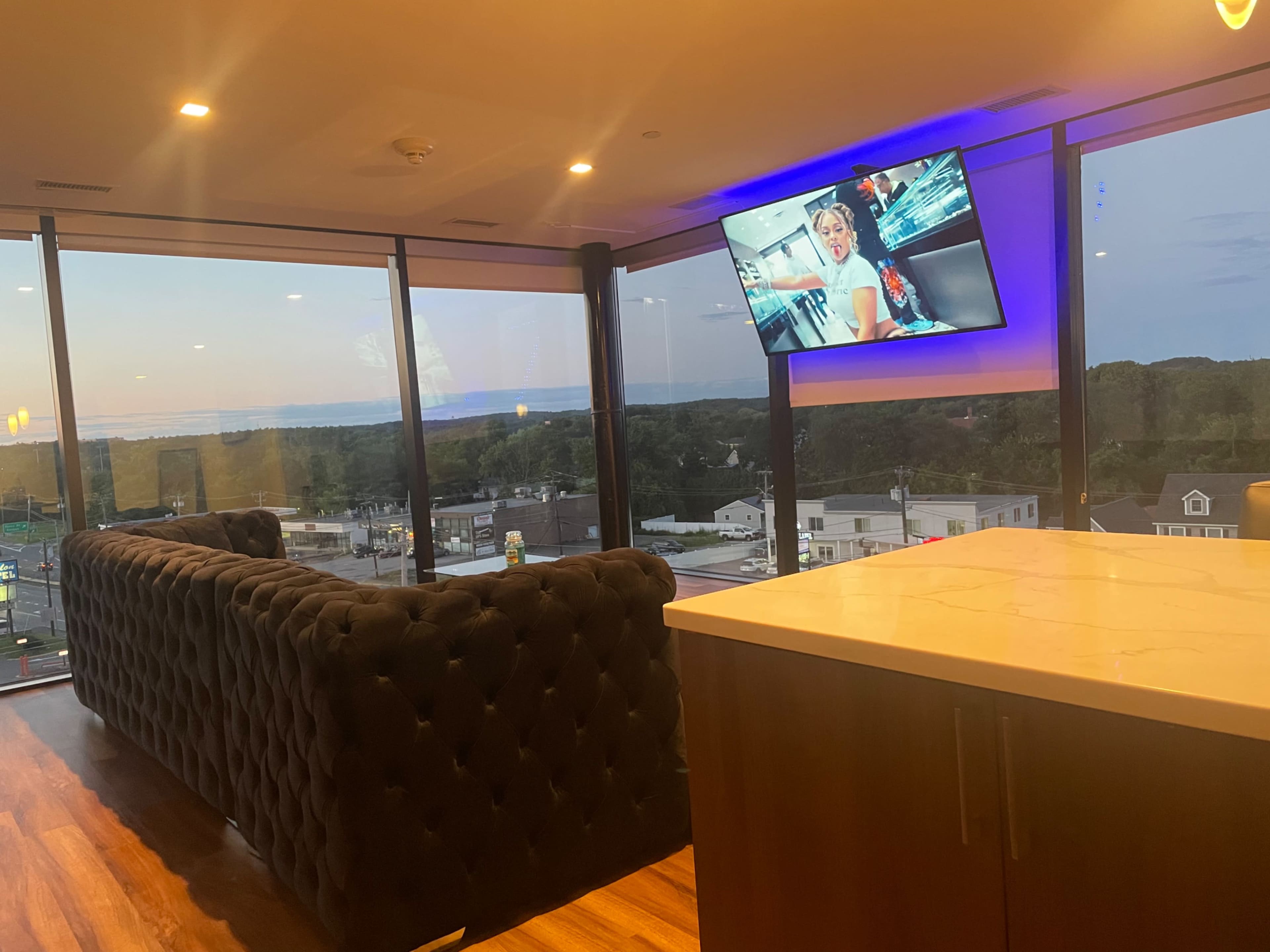 A modern living space features a dark sofa, a wall-mounted television displaying a program, and large windows offering a view of the landscape at sunset.