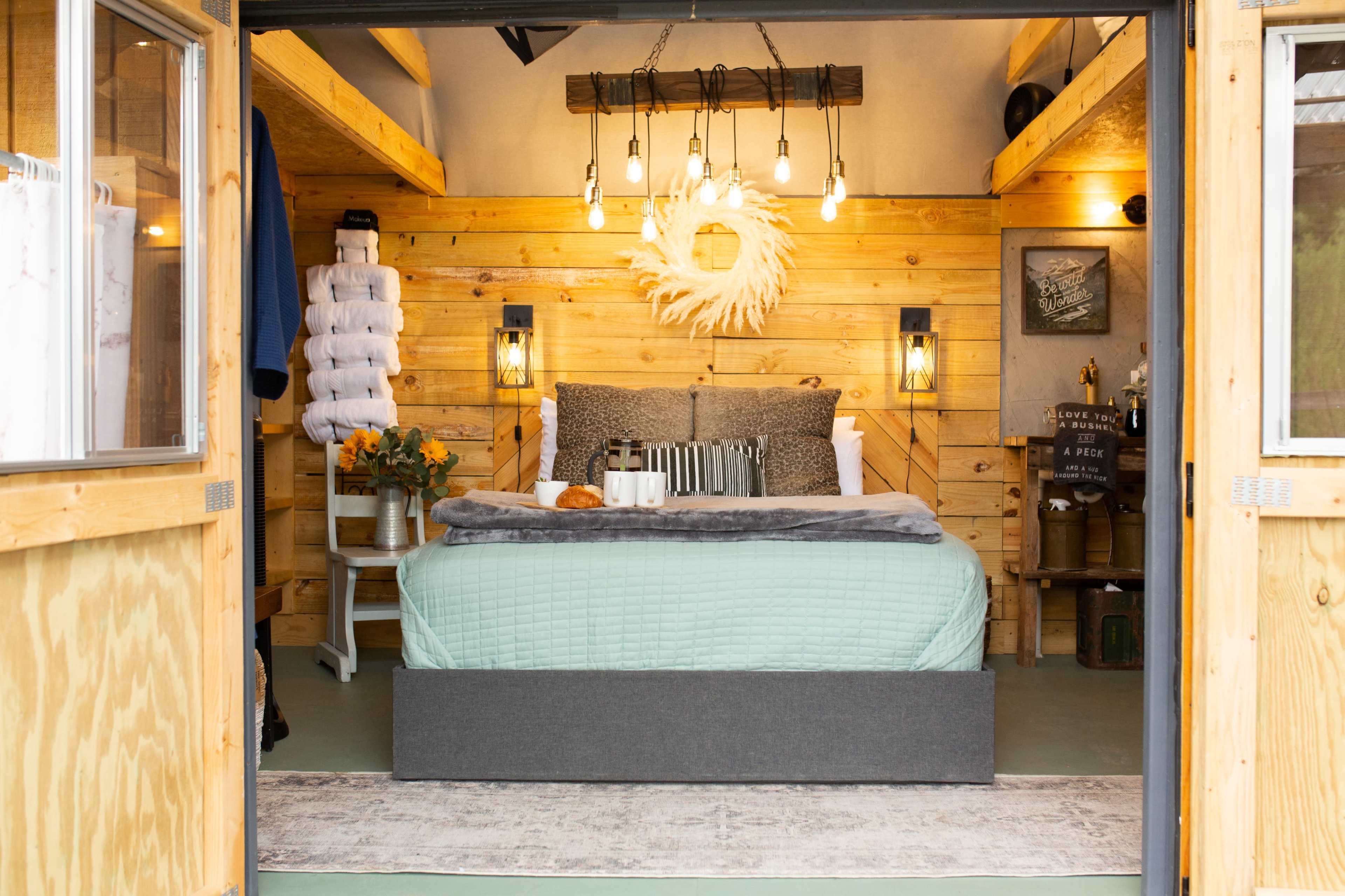 A cozy cabin interior features a neatly made bed with a light green quilt, wooden walls, a decorative chandelier, and rustic furnishings.