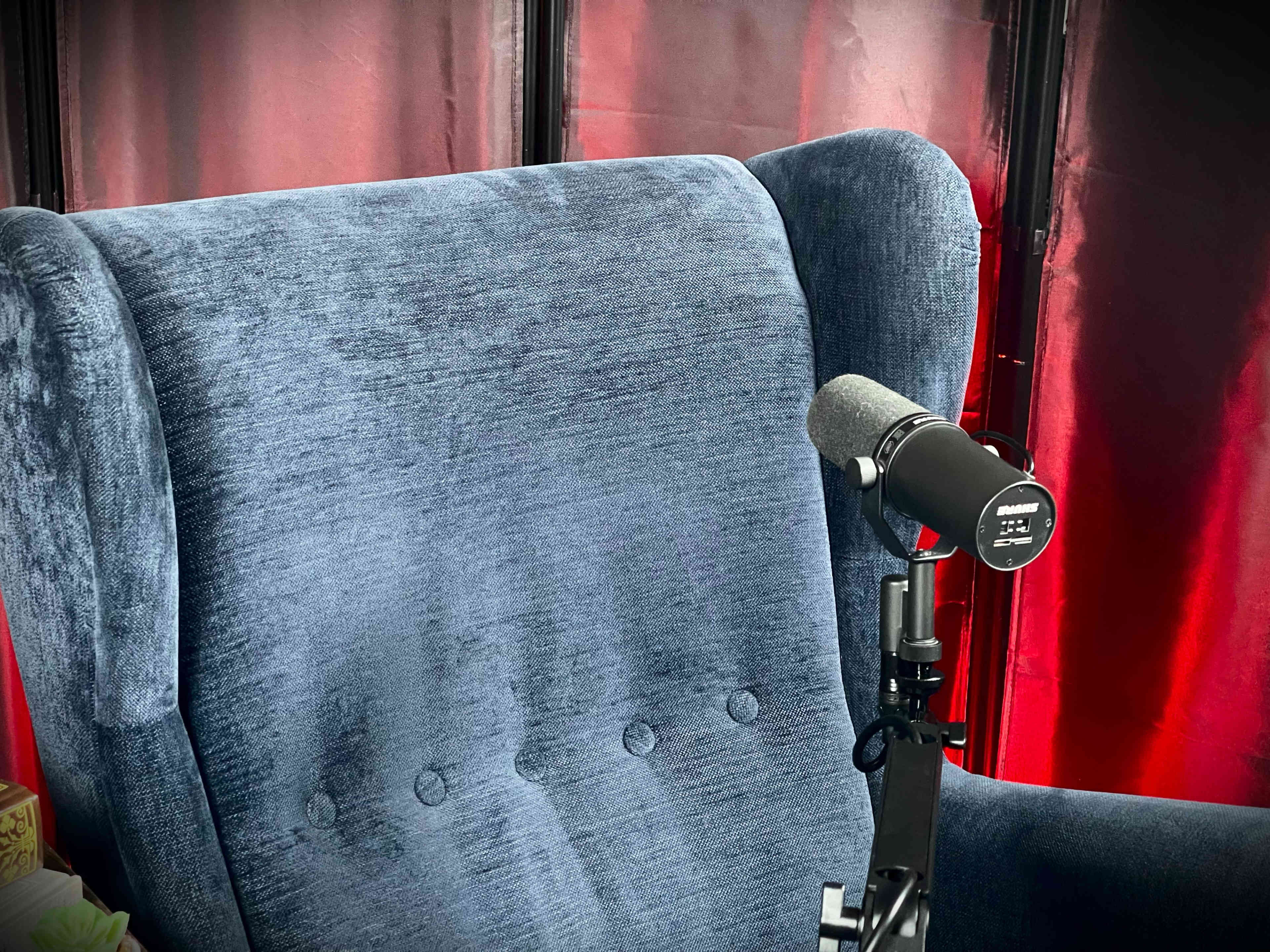 The image shows a blue upholstered chair with a microphone attached to a stand in front of it, set against a backdrop of red curtains.