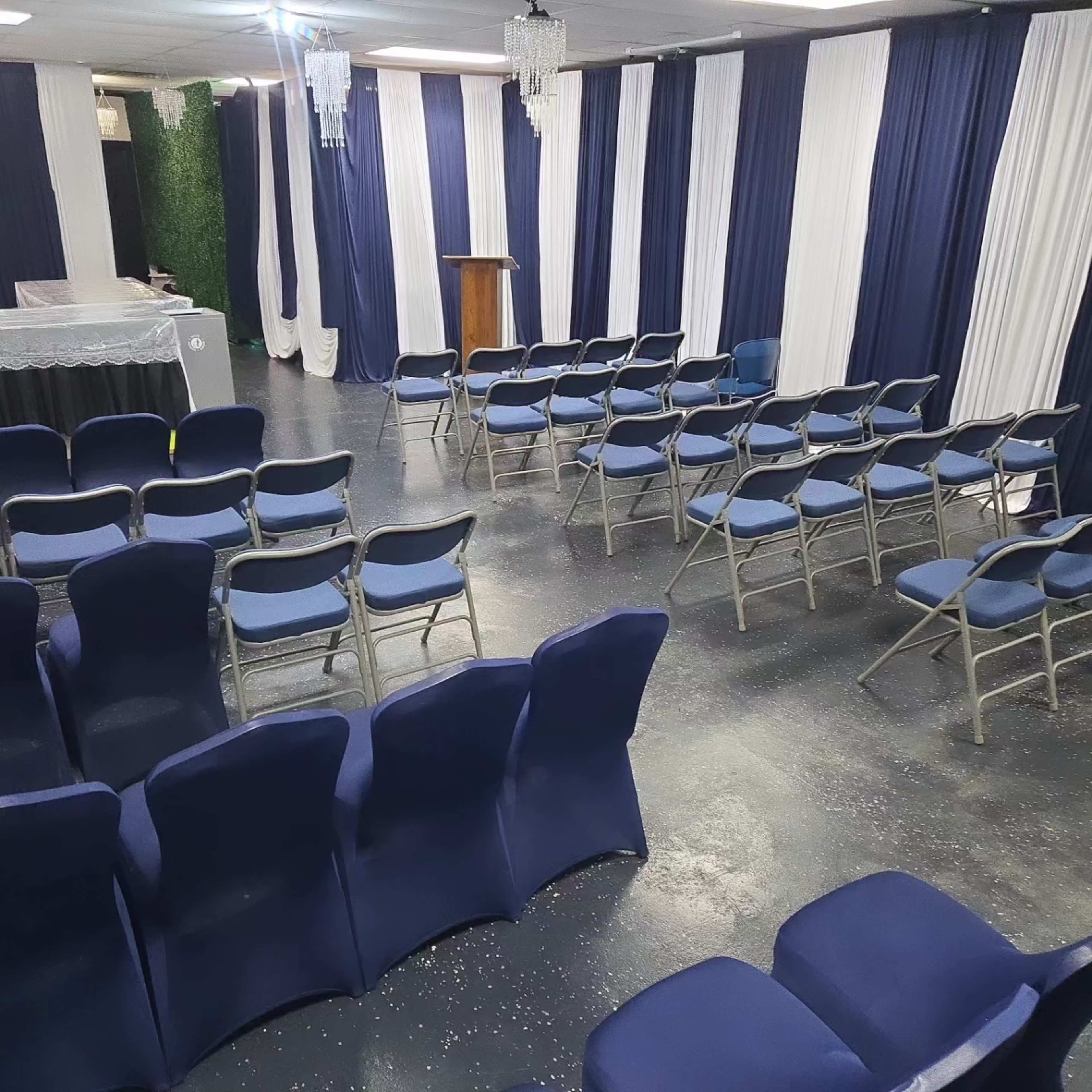 The image shows a small, neatly arranged event space with blue and white decor, featuring rows of blue chairs facing a wooden podium at the front.