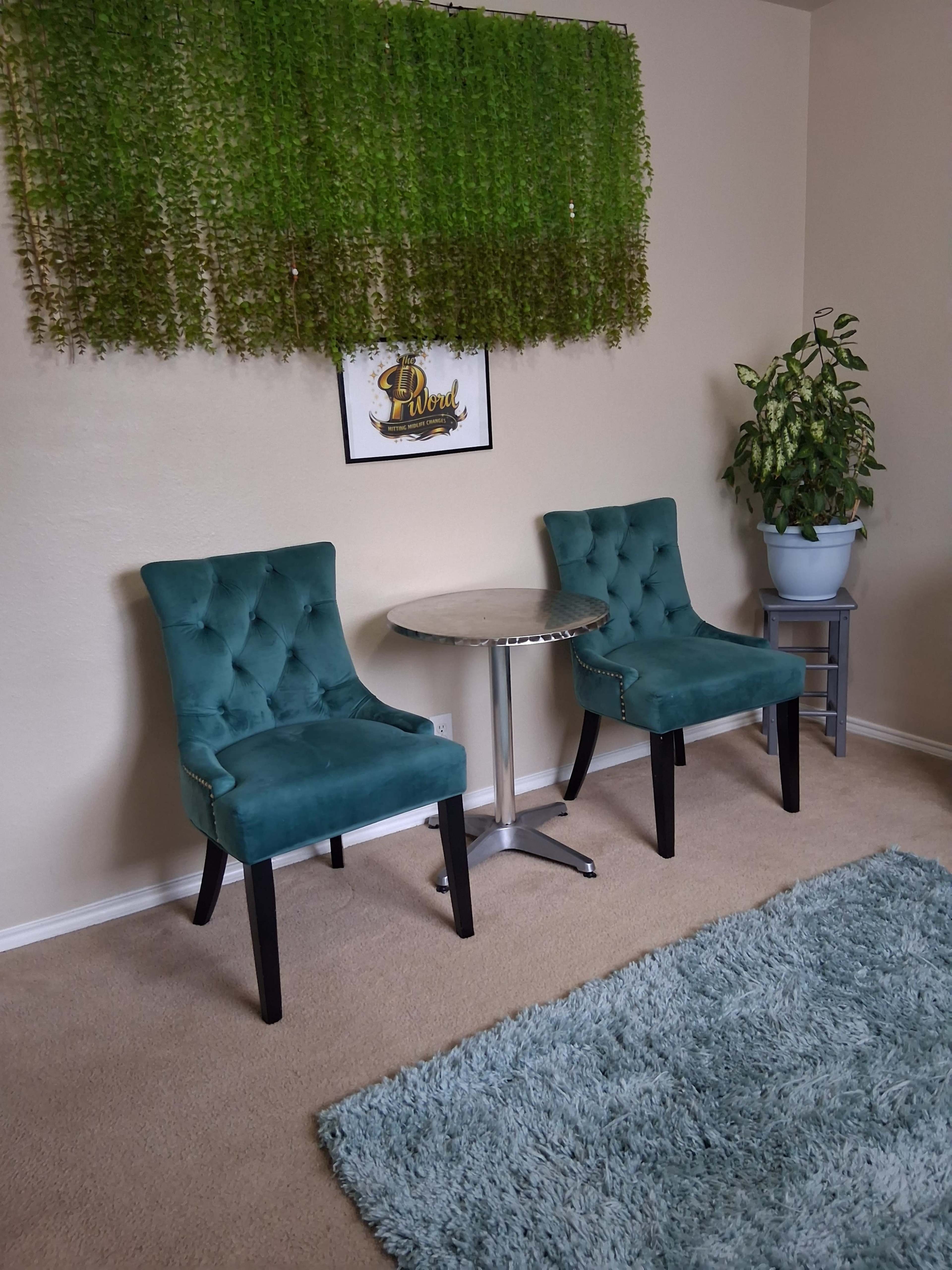 Two teal chairs are positioned around a small circular table, with a plant on a stand and a green wall hanging in the background.