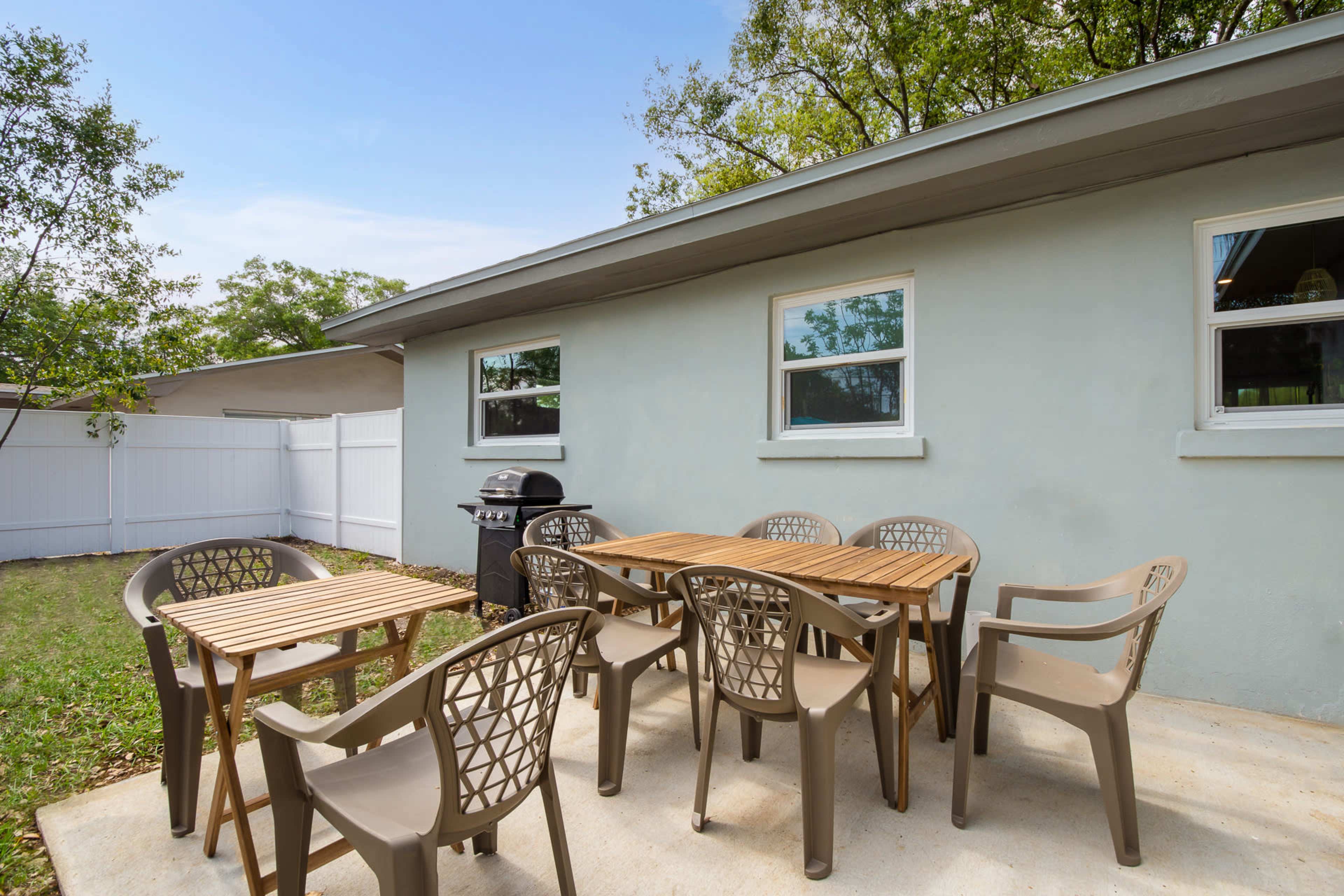 A patio features a wooden dining table surrounded by multiple chairs, with a grill and a fenced yard in the background.