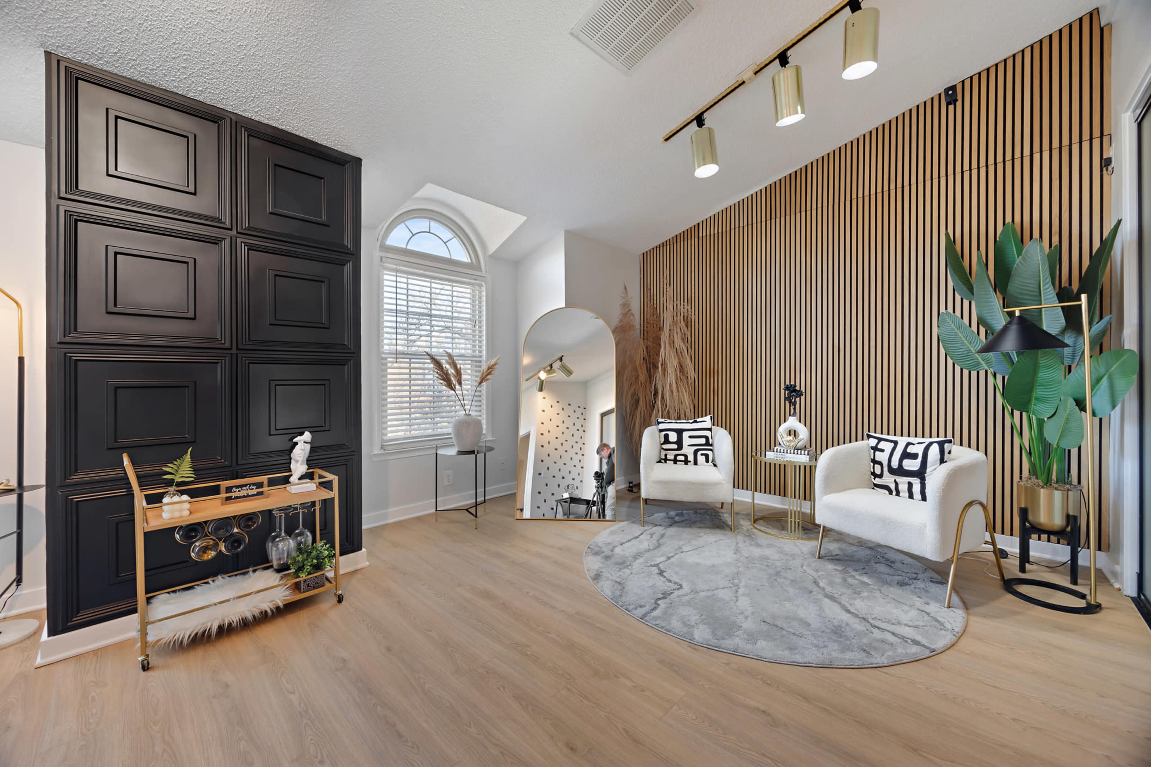 The image shows a modern living space featuring a stylish seating area with two armchairs, a round rug, a decorative wall with wood paneling, and a small bar cart.