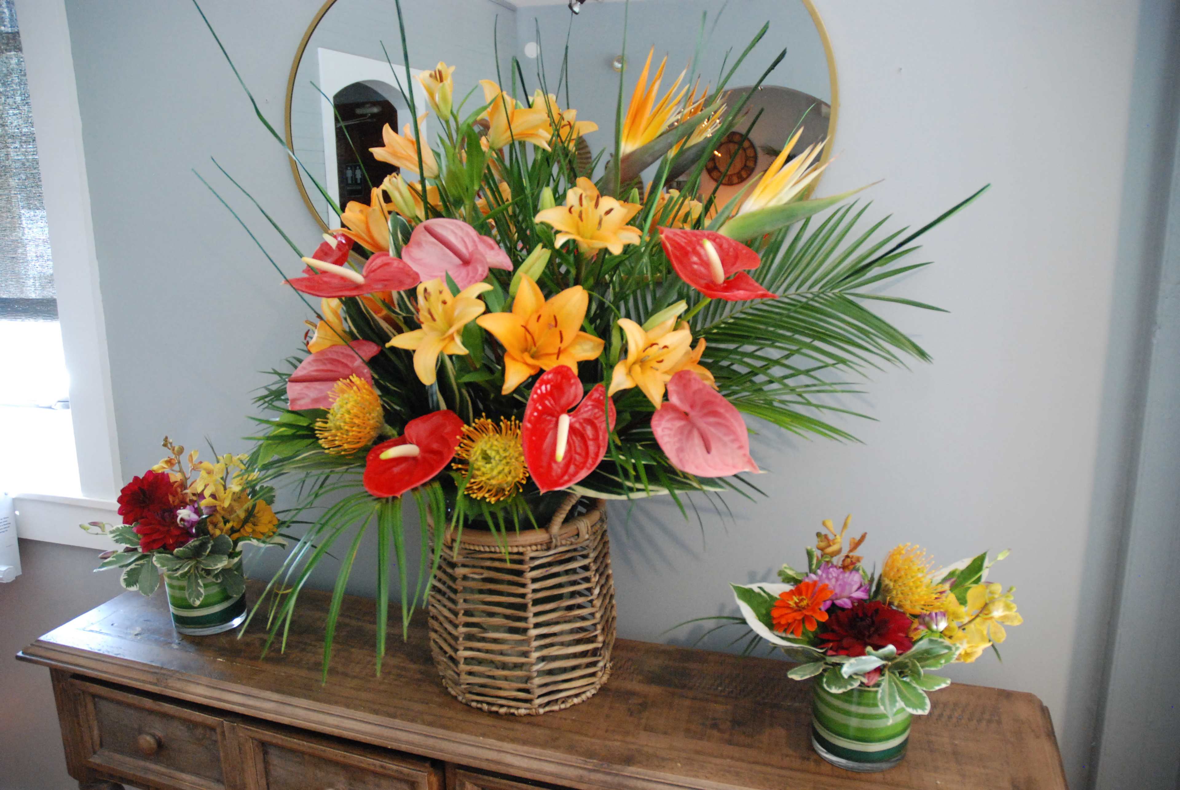 A large floral arrangement with tropical flowers and greenery is placed in a woven basket on a wooden table, accompanied by two smaller flower vases on either side.
