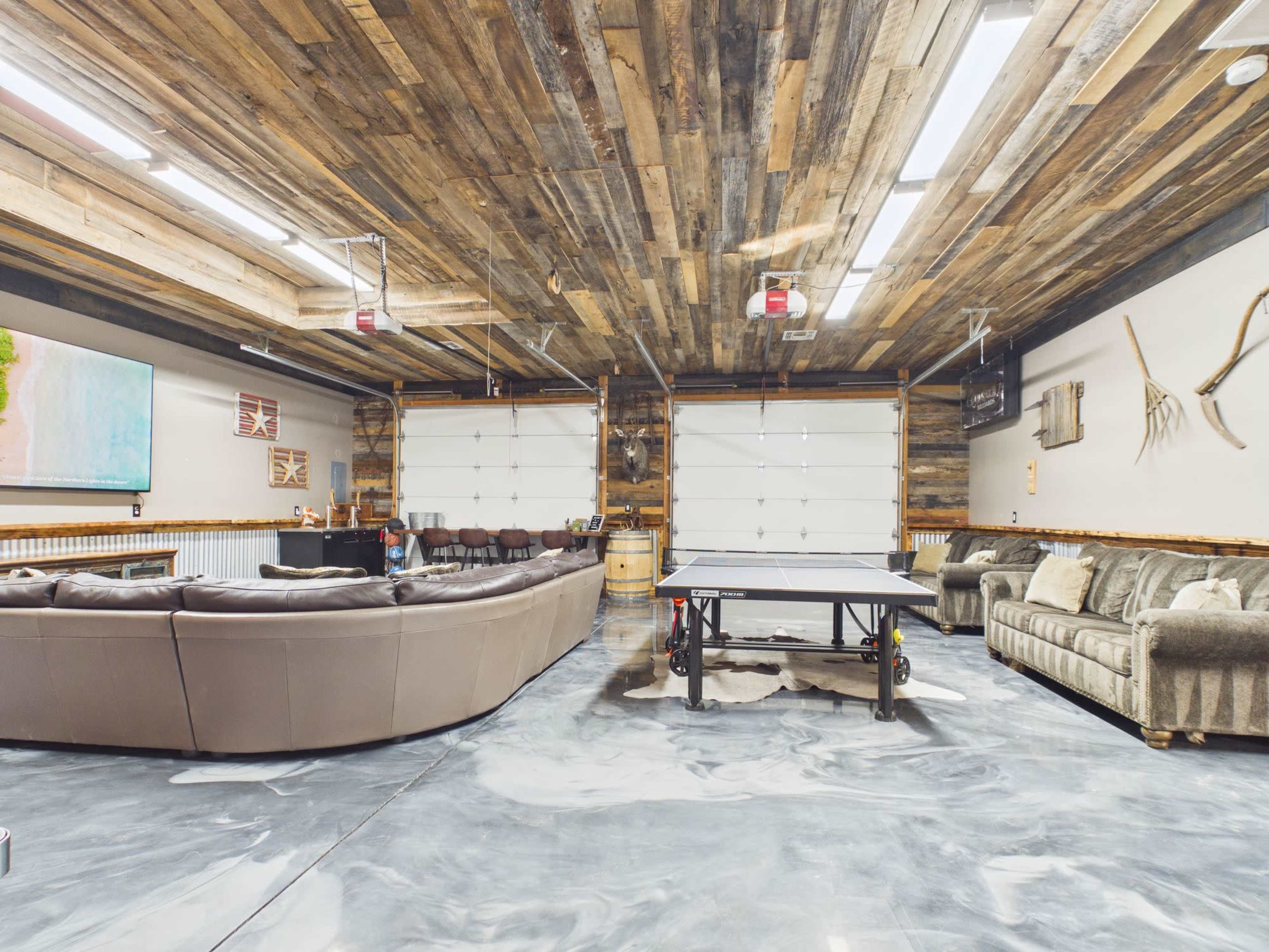 The image shows a spacious garage converted into a recreational room, featuring a ping pong table surrounded by a sectional couch and a sofa, with wooden paneling on the ceiling and walls.