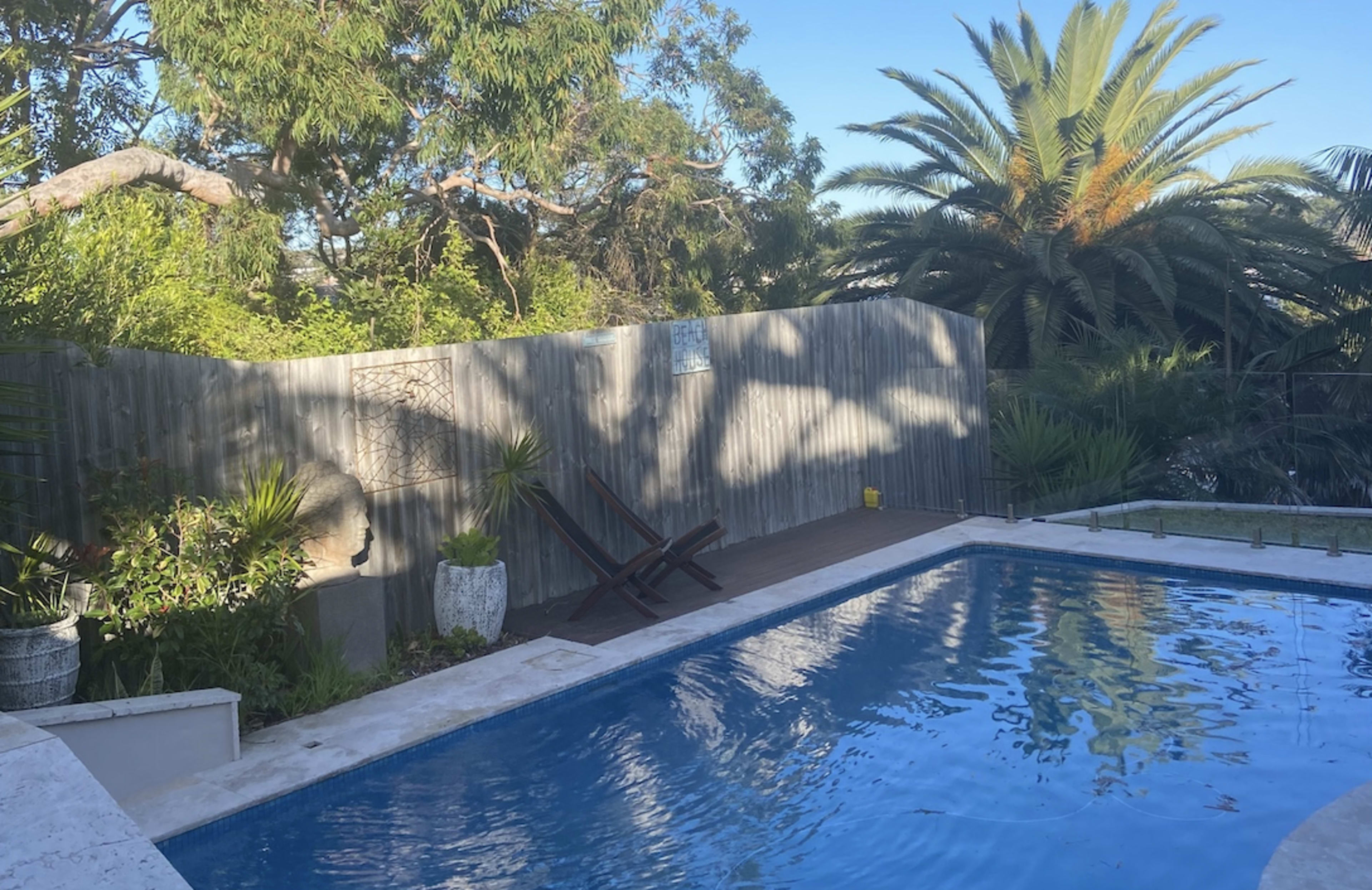 A swimming pool is surrounded by greenery and a wooden deck with a pair of lounge chairs.