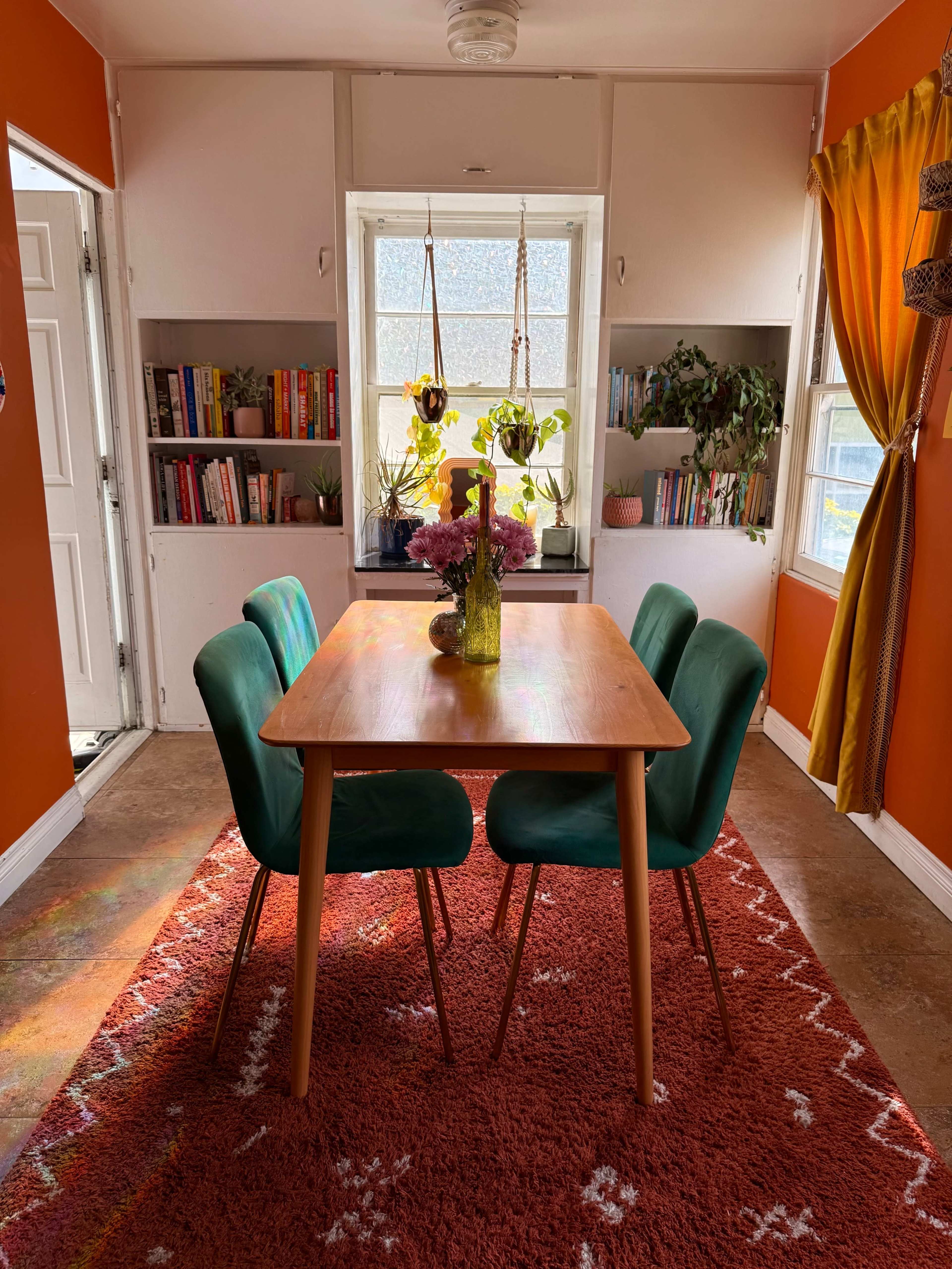 A wooden table with four green chairs is set in a brightly colored room with orange walls and a rug, surrounded by bookshelves and plants.