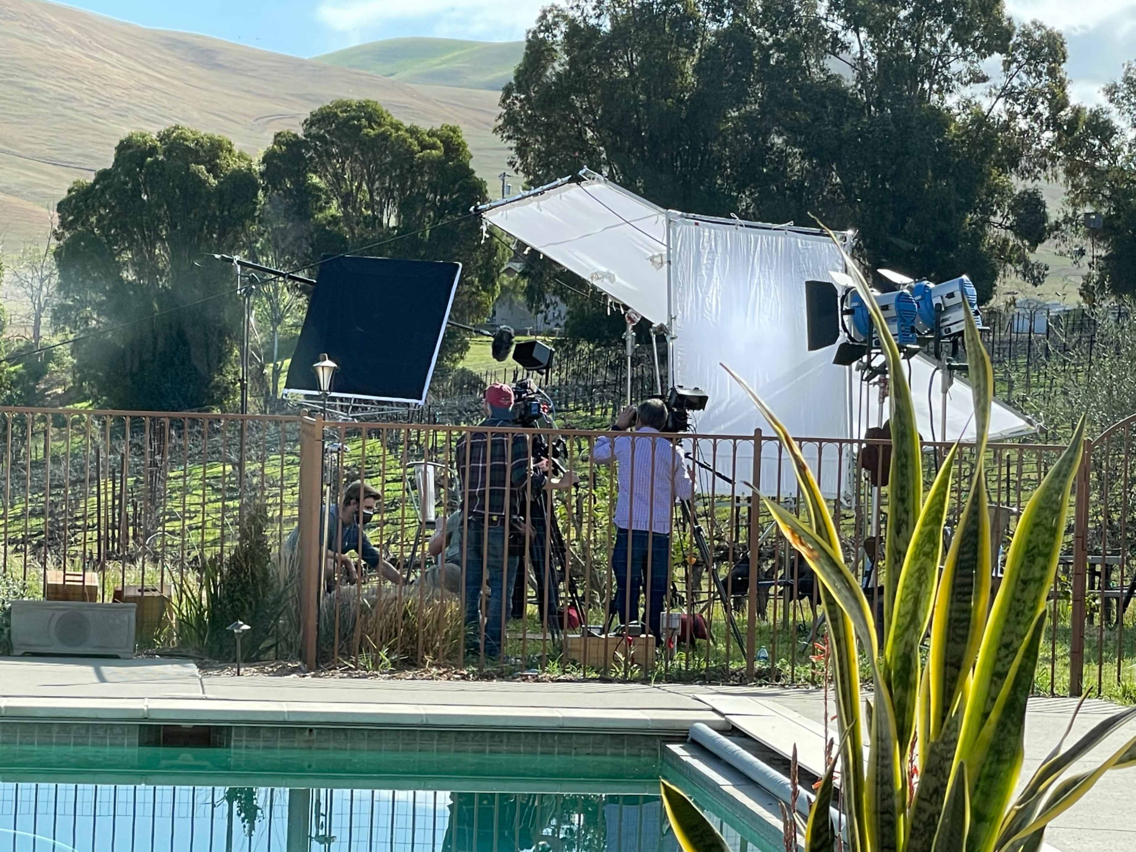 A film crew is set up near a swimming pool on a property with vineyards in the background, featuring large lighting equipment and cameras.