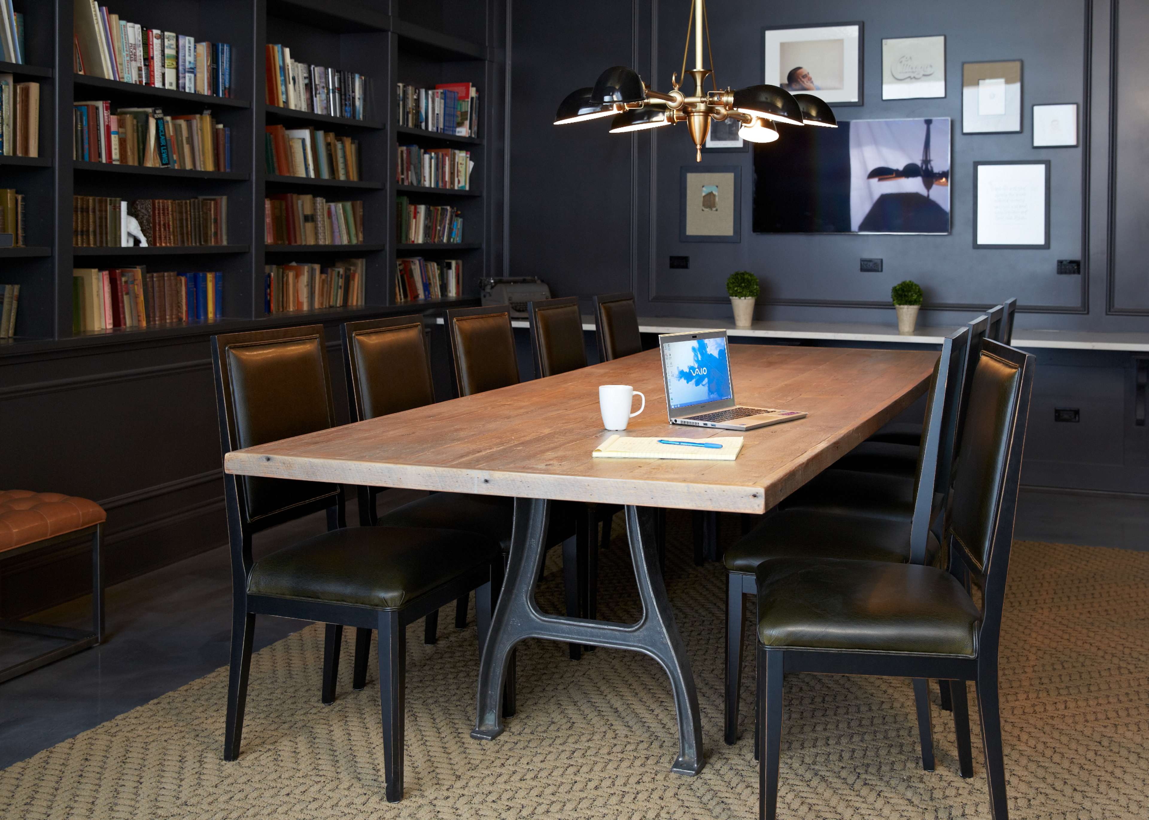 A long wooden table with eight black chairs is set in a room with bookshelves and a large wall-mounted screen.