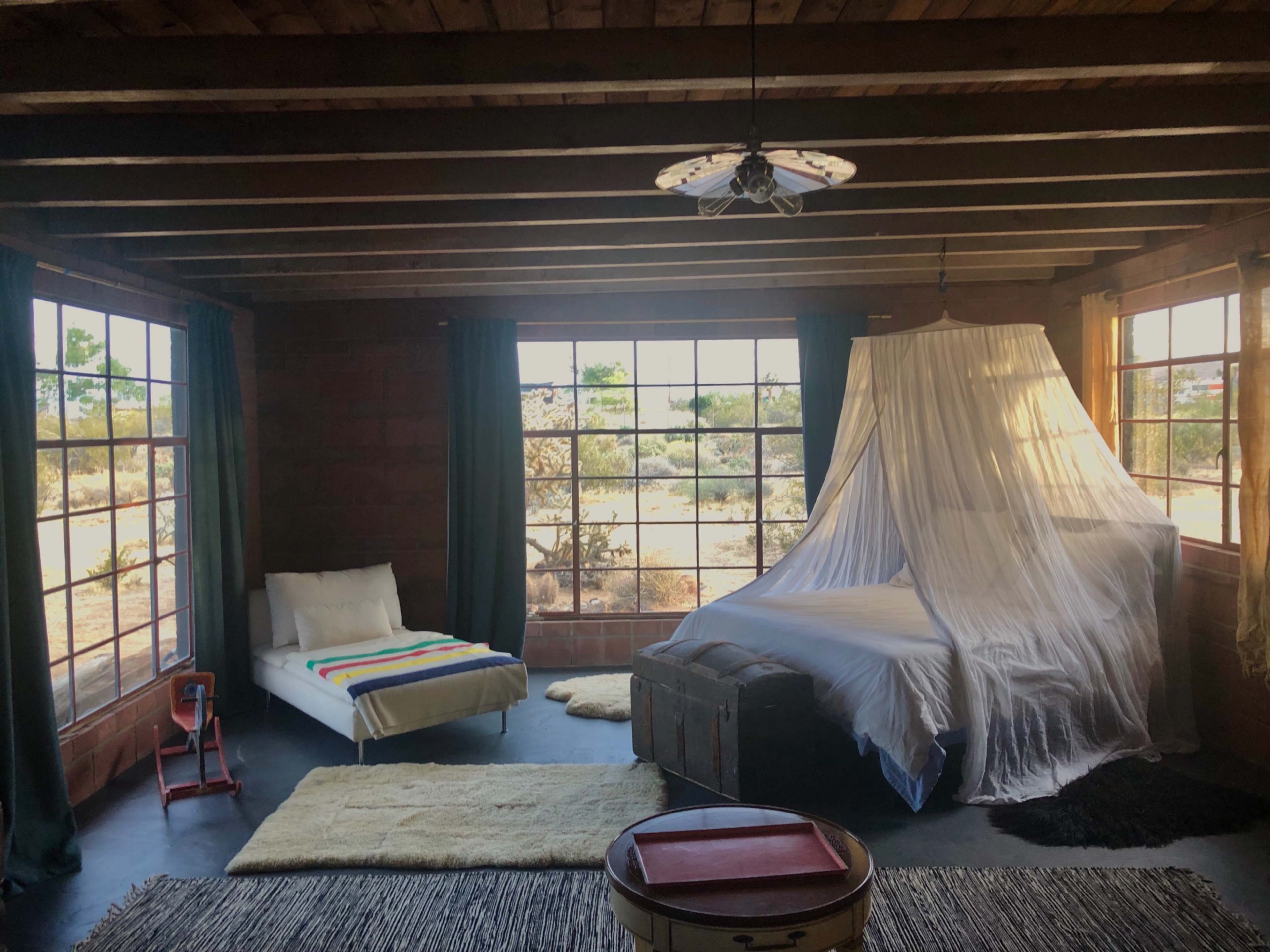 A rustic bedroom features a bed with a canopy, a white lounge chair, and large windows overlooking a desert landscape.