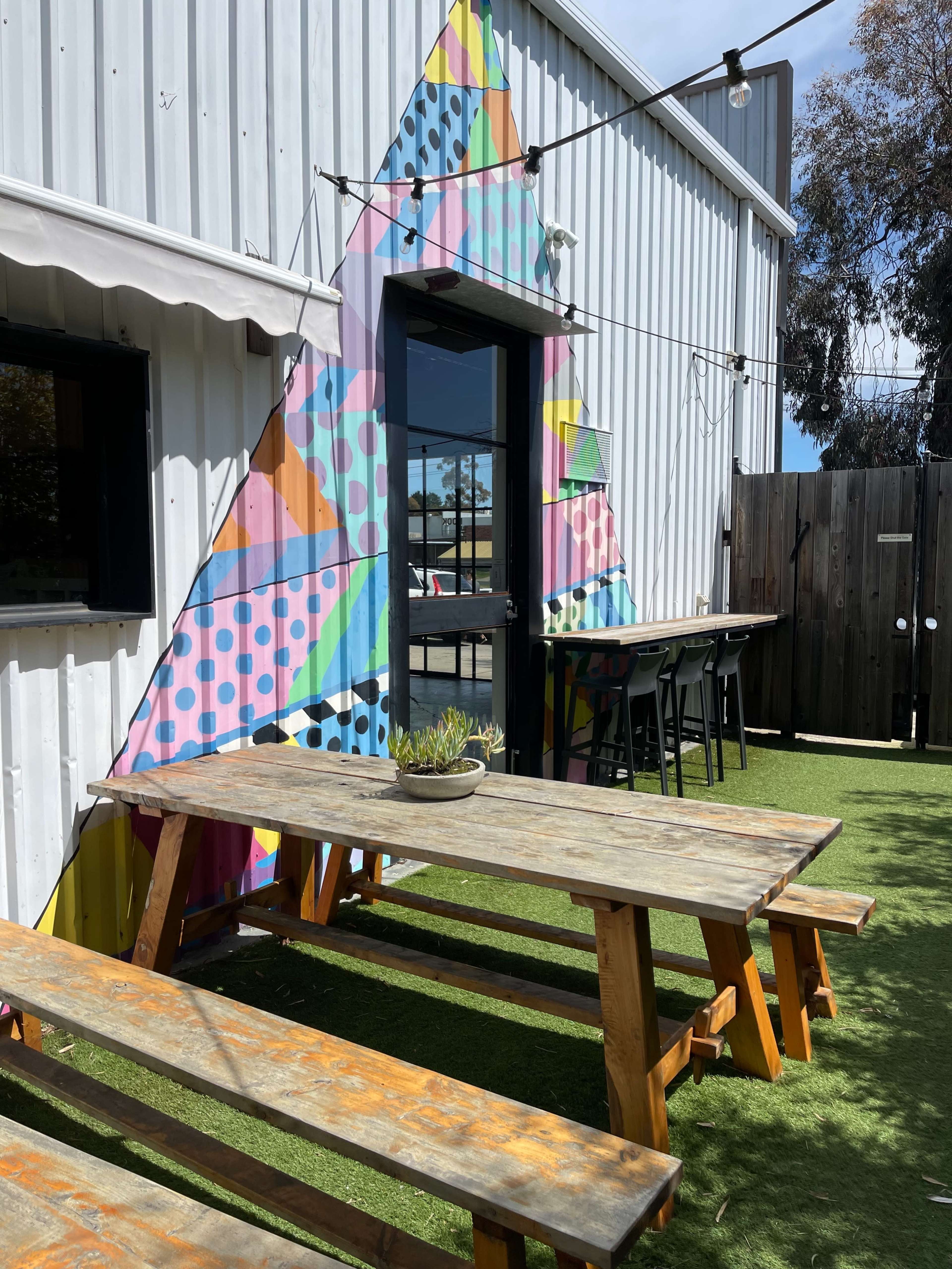 A colorful triangular mural adorns the entrance of a building, with outdoor wooden picnic tables and a green grass-like surface nearby.
