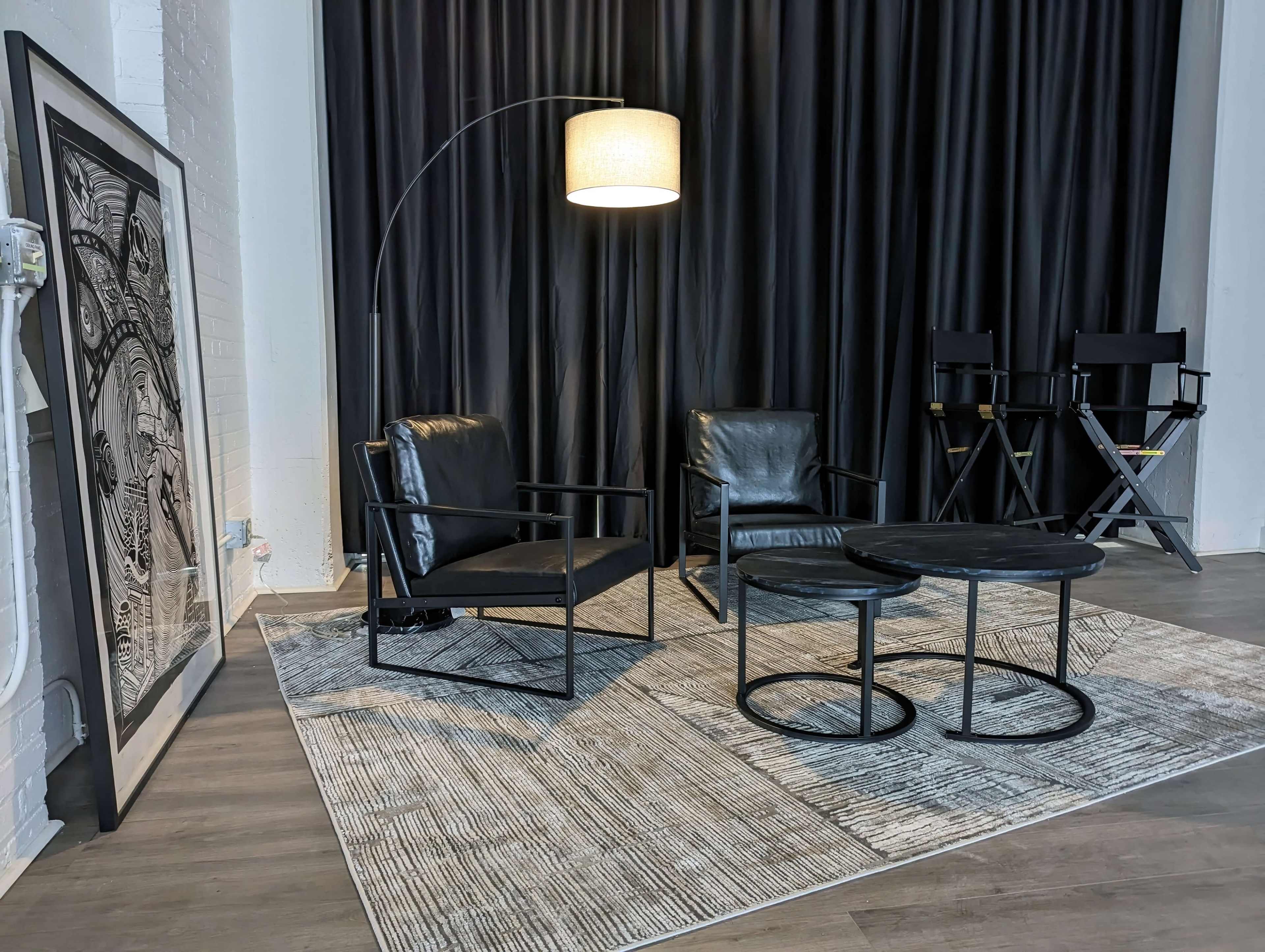 The image features a seating arrangement with two black leather chairs, a circular side table, and an arc floor lamp, set against a backdrop of dark curtains and a large framed artwork.