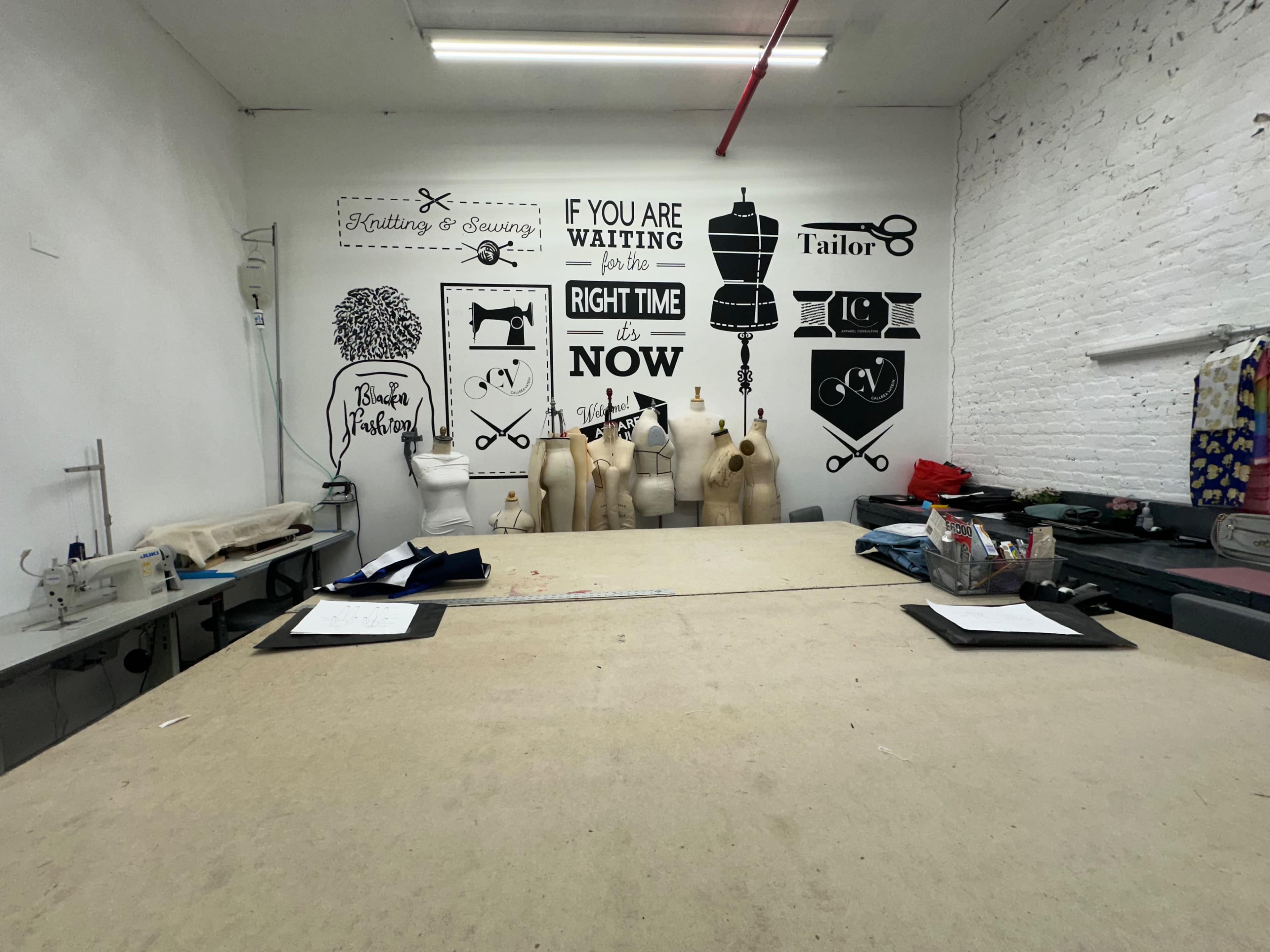 A sewing studio with a large cutting table at the center and a wall decorated with sewing-related graphics and text.