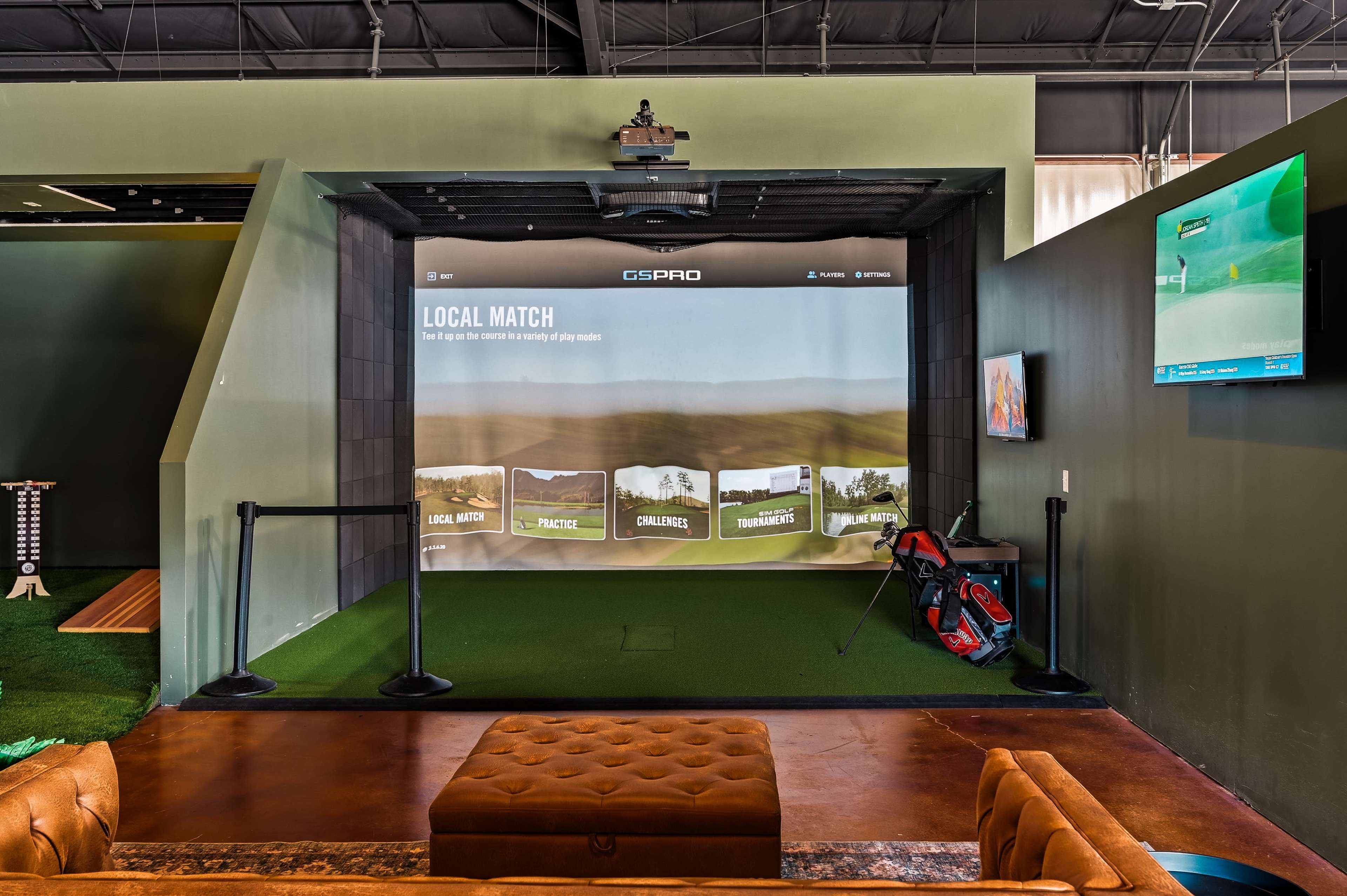 The image shows an indoor golf simulator setup with a large screen displaying a golf course scene, surrounded by green turf and golf bags.