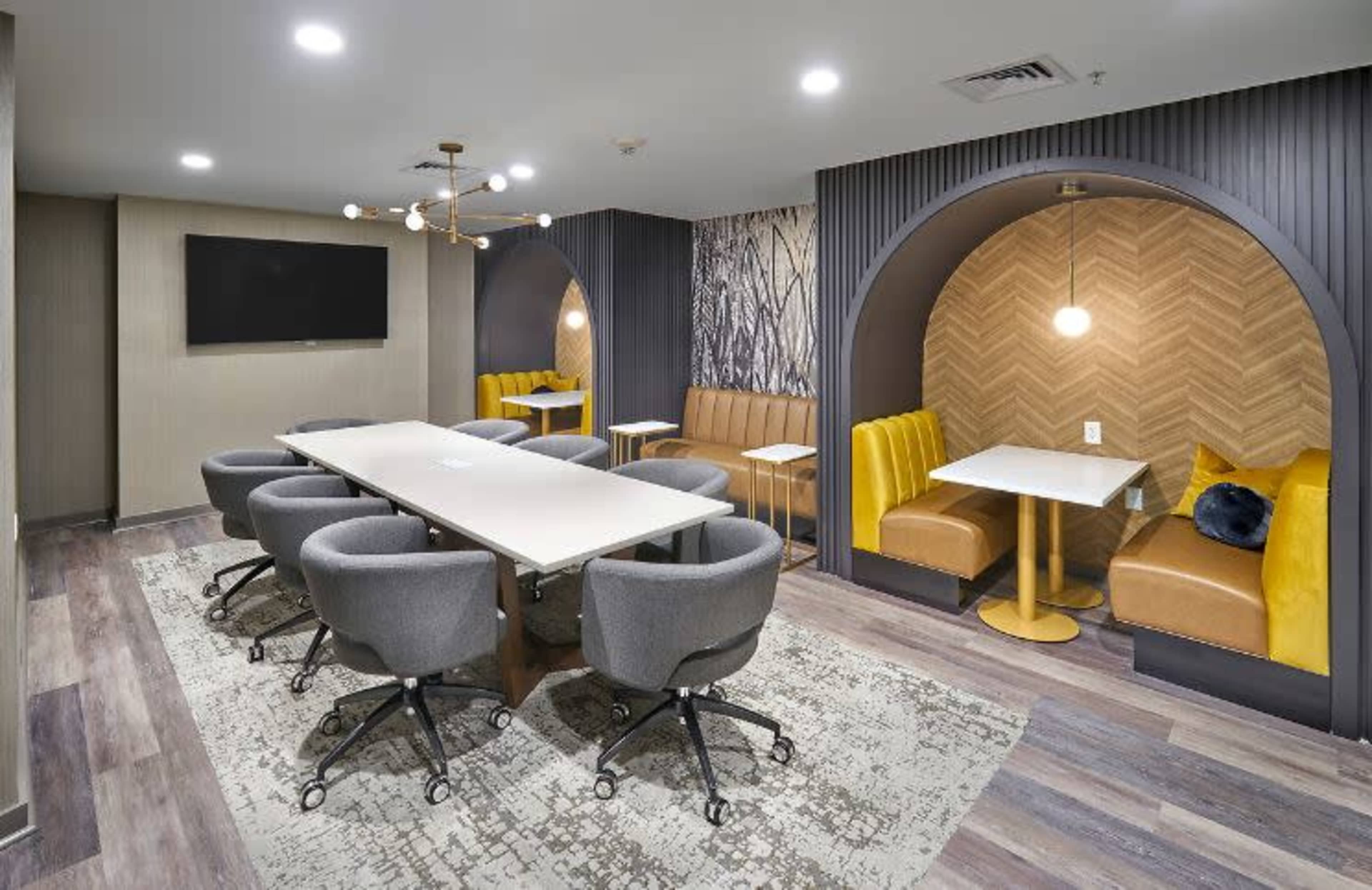 A modern meeting room with a long table surrounded by chairs, two booth seating areas, and a wall-mounted television.
