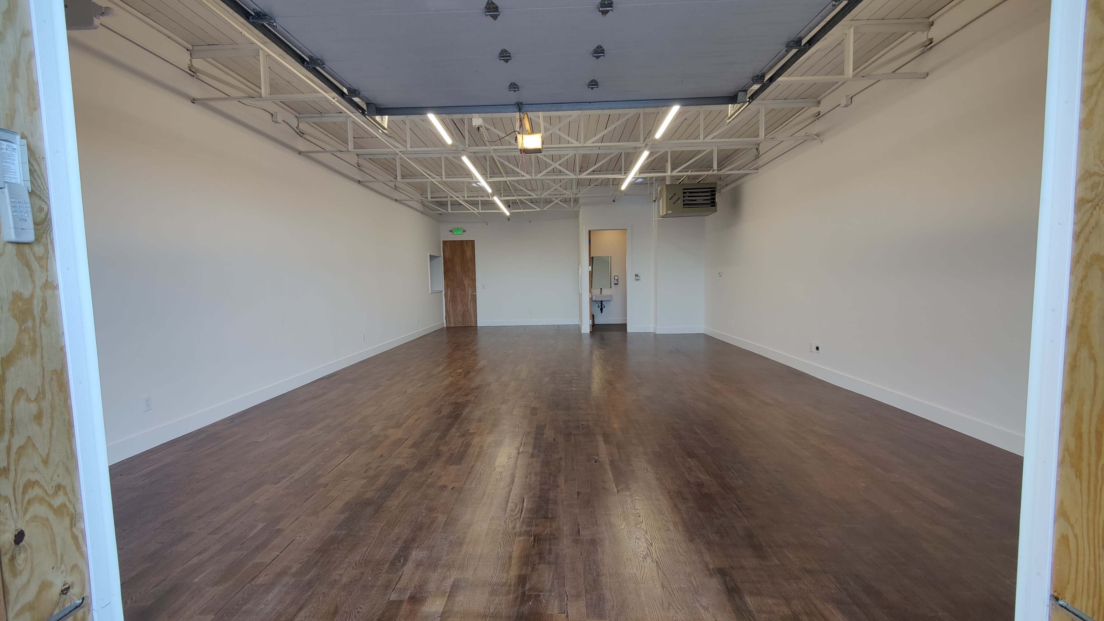 The image shows a spacious, empty room with hardwood floors and white walls, illuminated by overhead lighting.