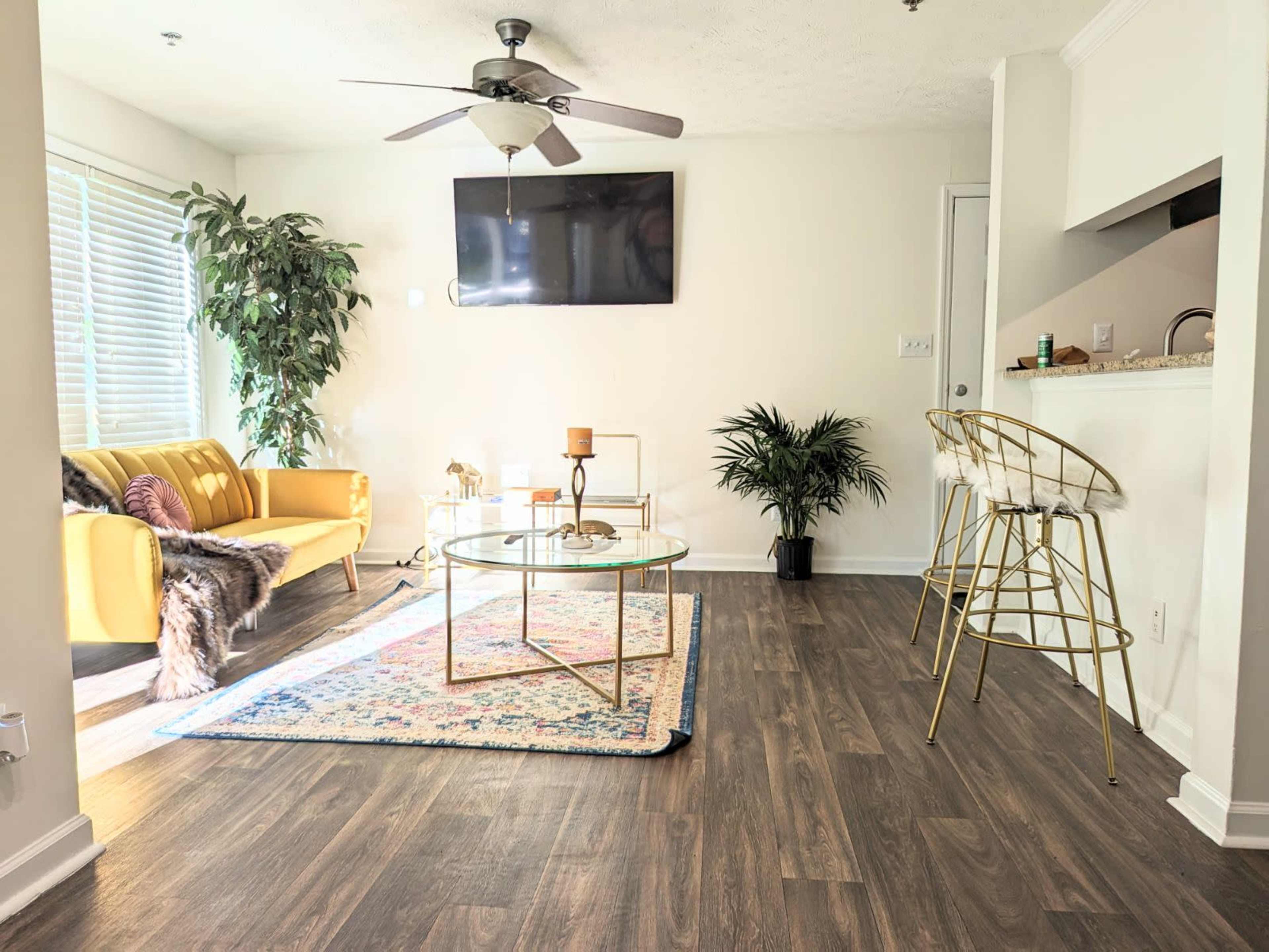 A bright living room featuring a yellow sofa, a glass coffee table on a colorful rug, and bar stools near a kitchen counter.