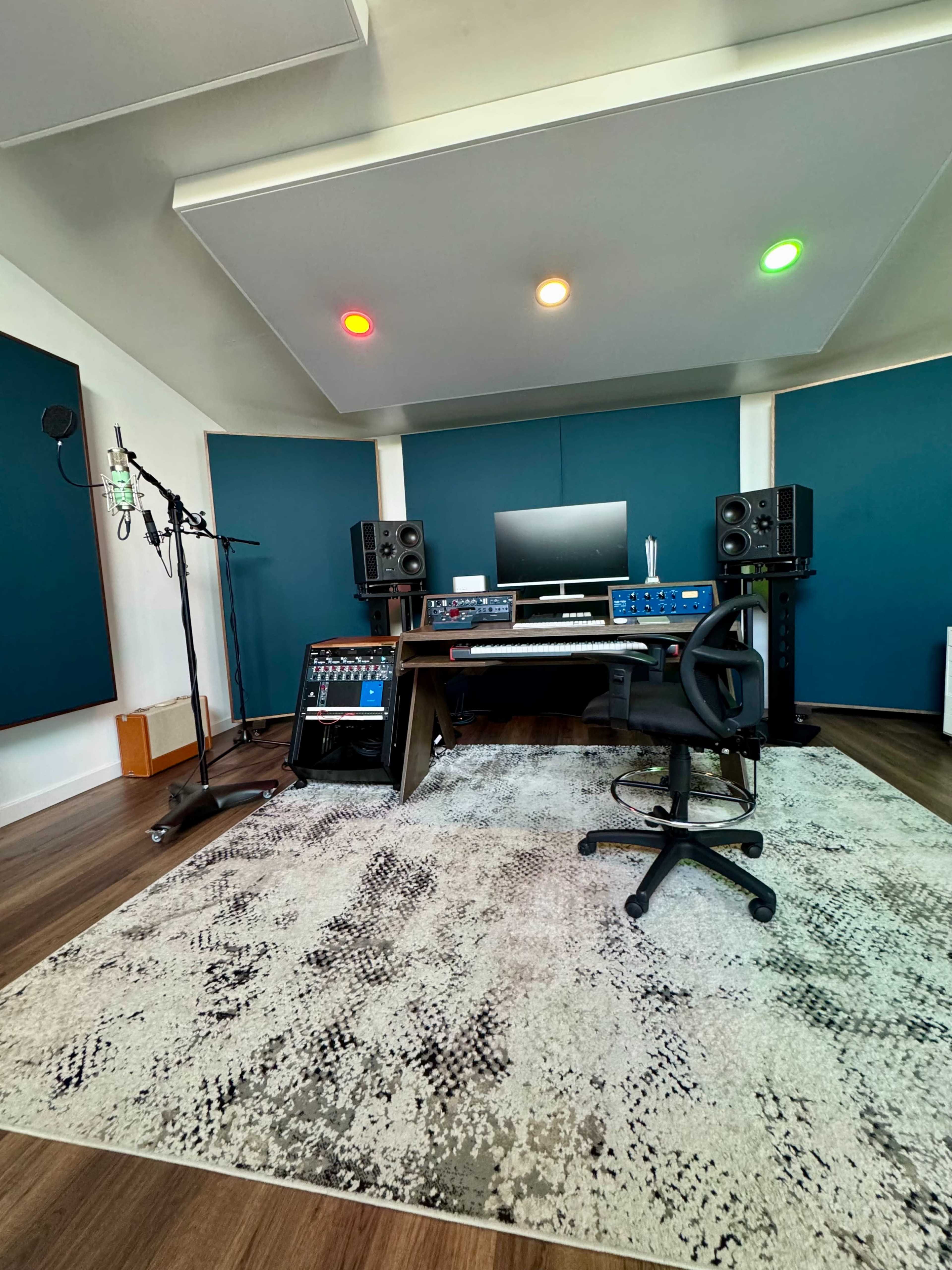 The image shows a modern recording studio with a computer, audio equipment, microphones, and a swivel chair set against teal soundproofing panels.