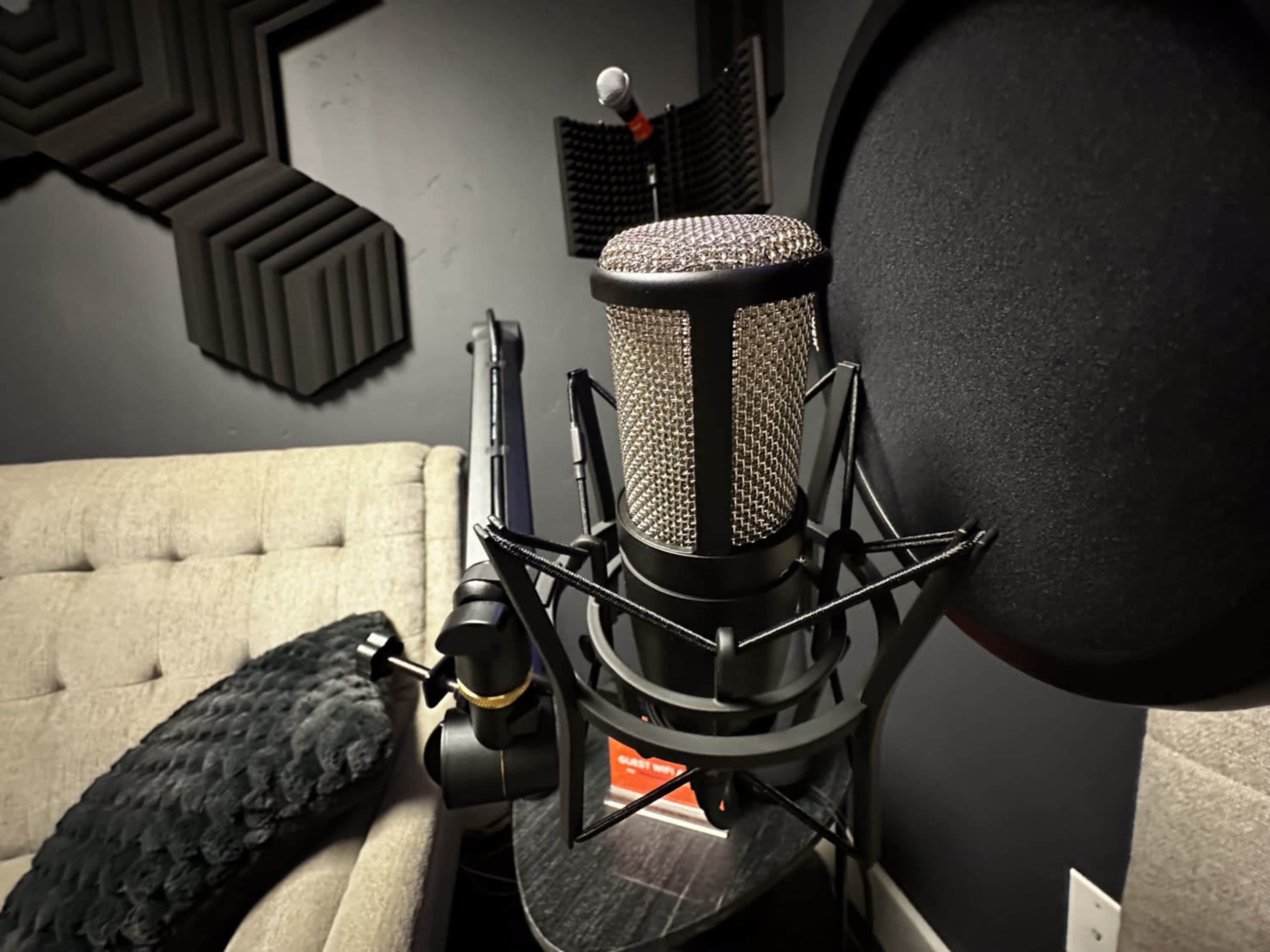 A microphone is positioned in front of a dark wall, next to a couch and soundproofing panels.