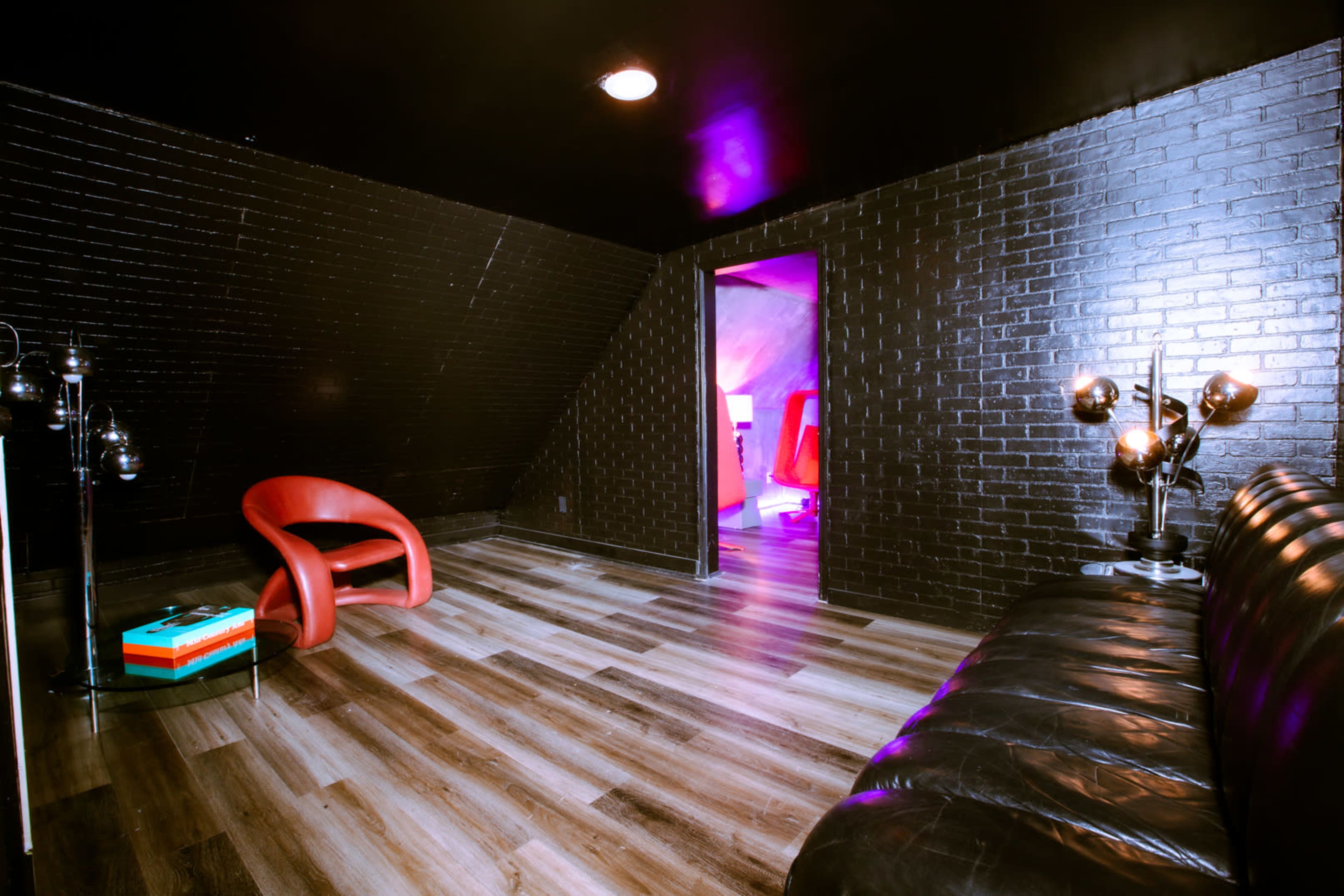 A dimly lit room with black brick walls, a leather sofa, a red chair, and a doorway leading to a brightly colored space.