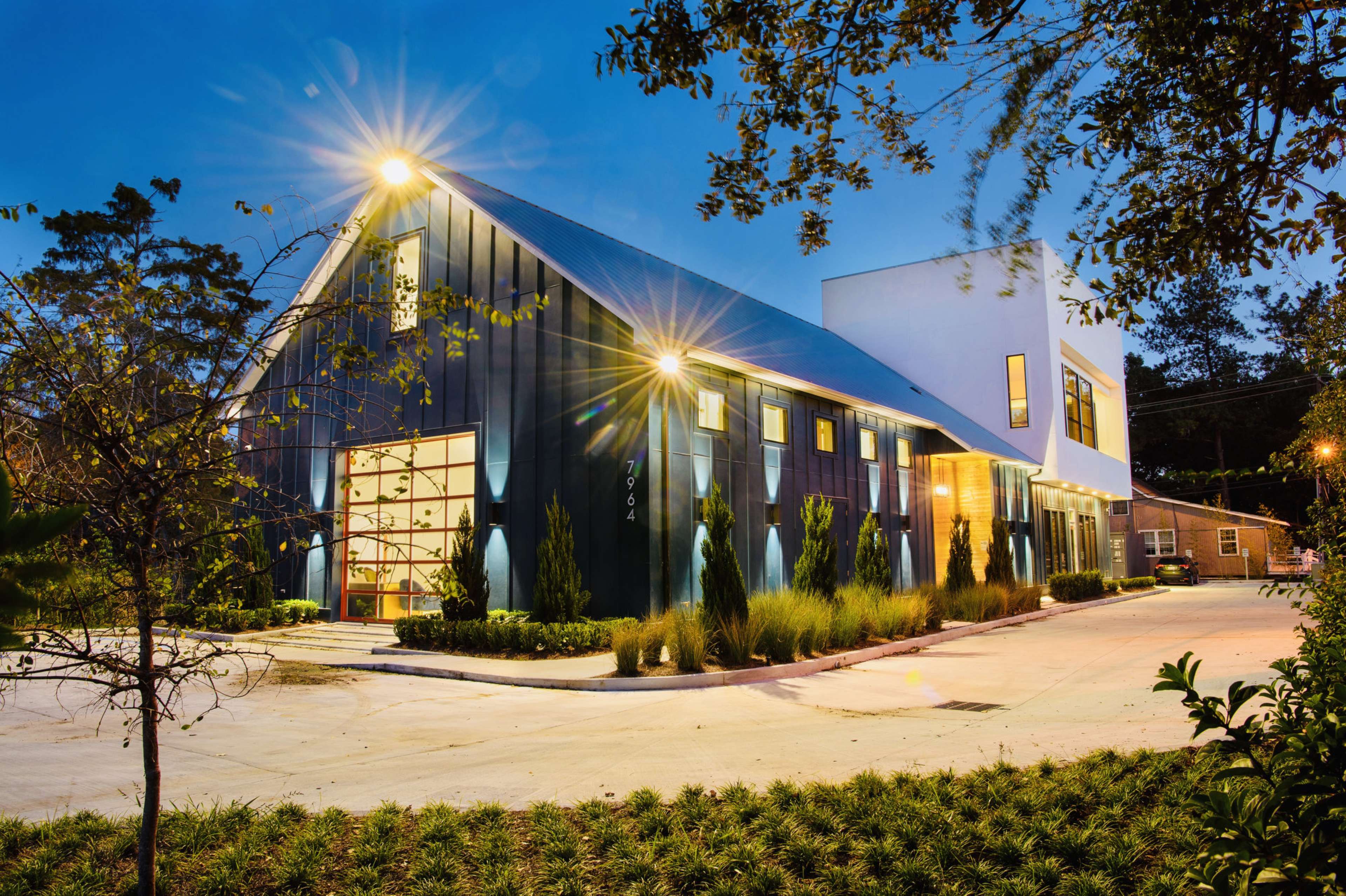 A modern building with a black exterior, large windows, and a white section, illuminated at dusk with landscaping and a driveway in the foreground.