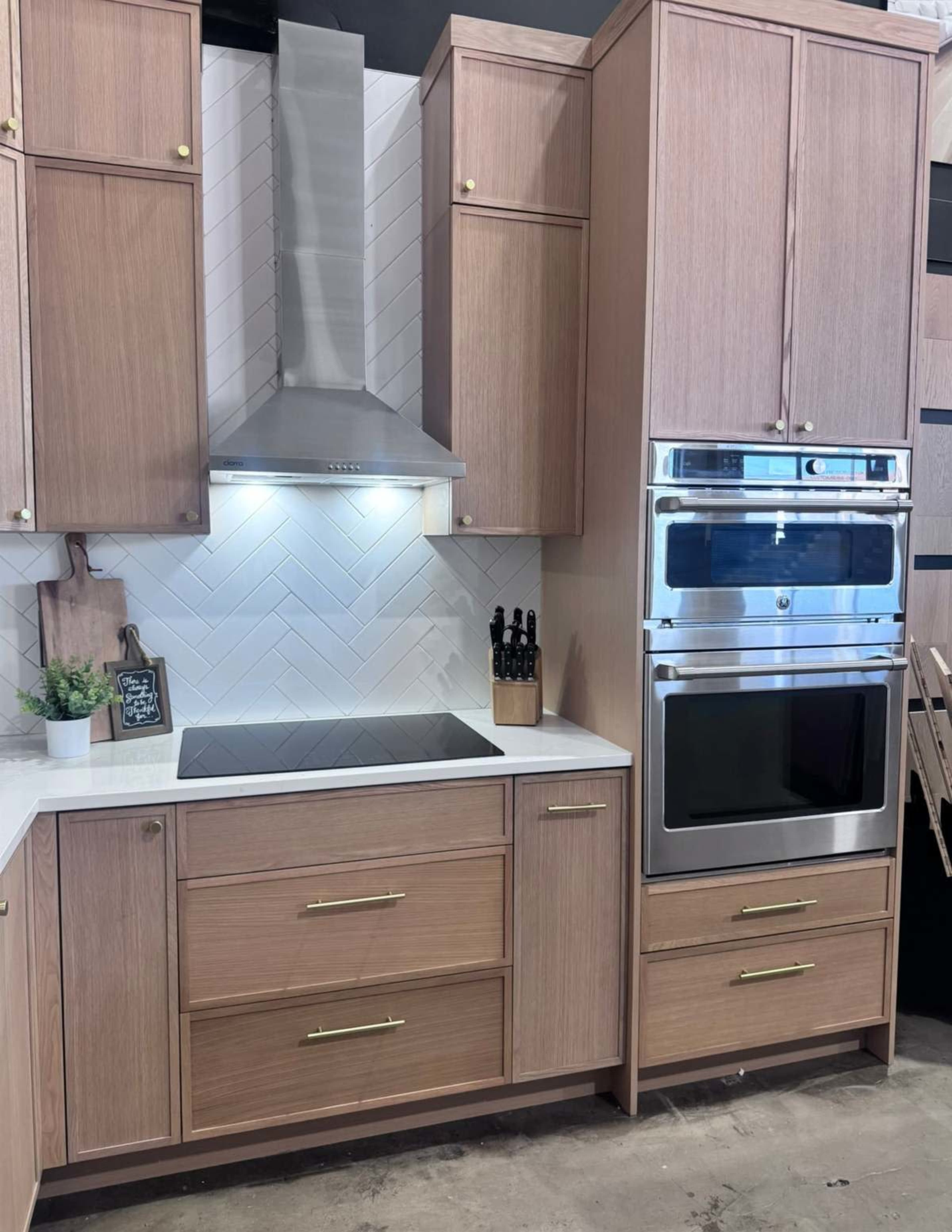 The image shows a modern kitchen setup featuring wooden cabinets, a stainless steel range hood, a built-in oven, and a herringbone tile backsplash.