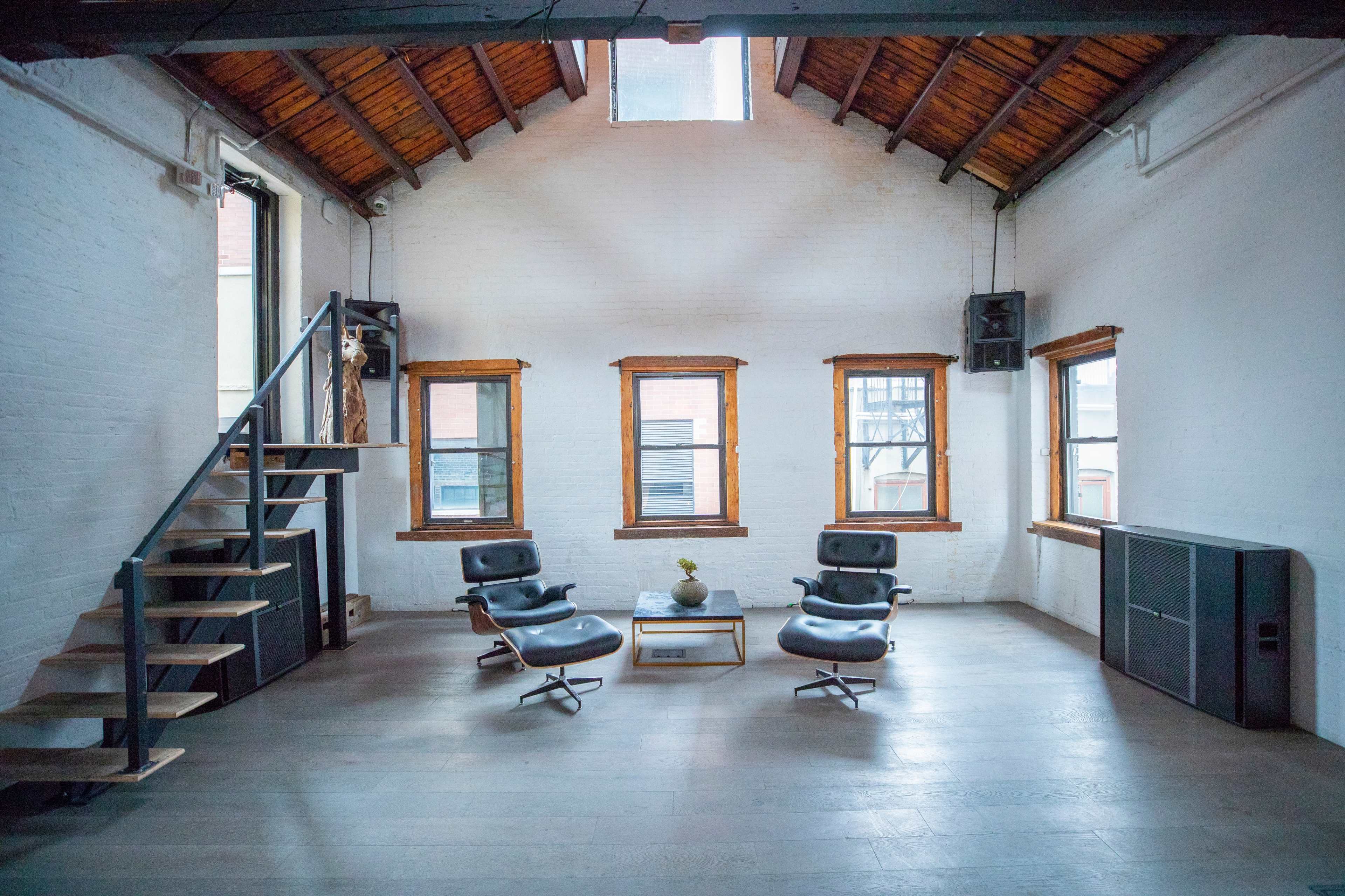 A spacious room with high ceilings, featuring two chairs, a table, and large windows, along with a staircase leading to an upper level.
