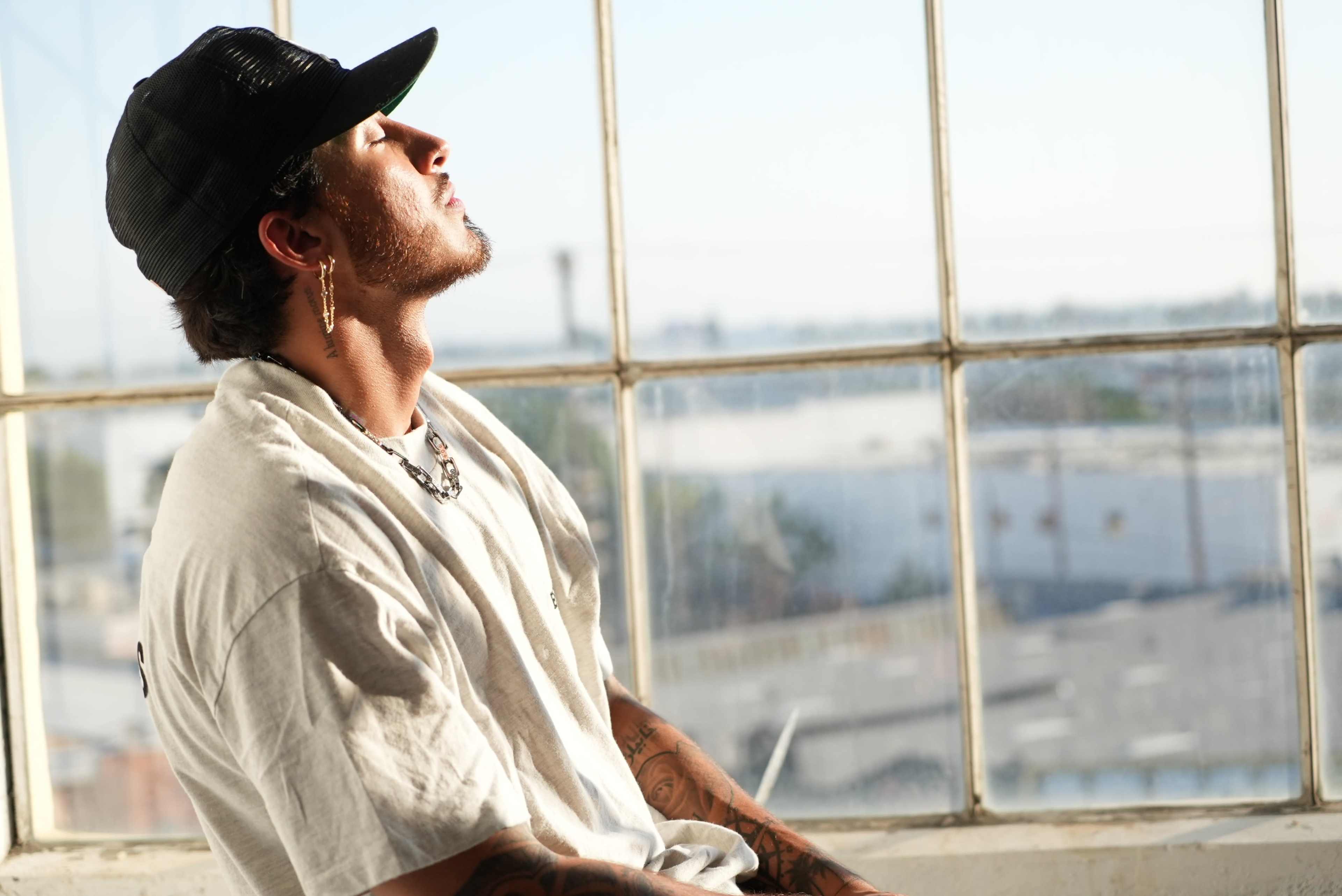 A man wearing a black cap and a white t-shirt sits by a large window, gazing upwards with his eyes closed.