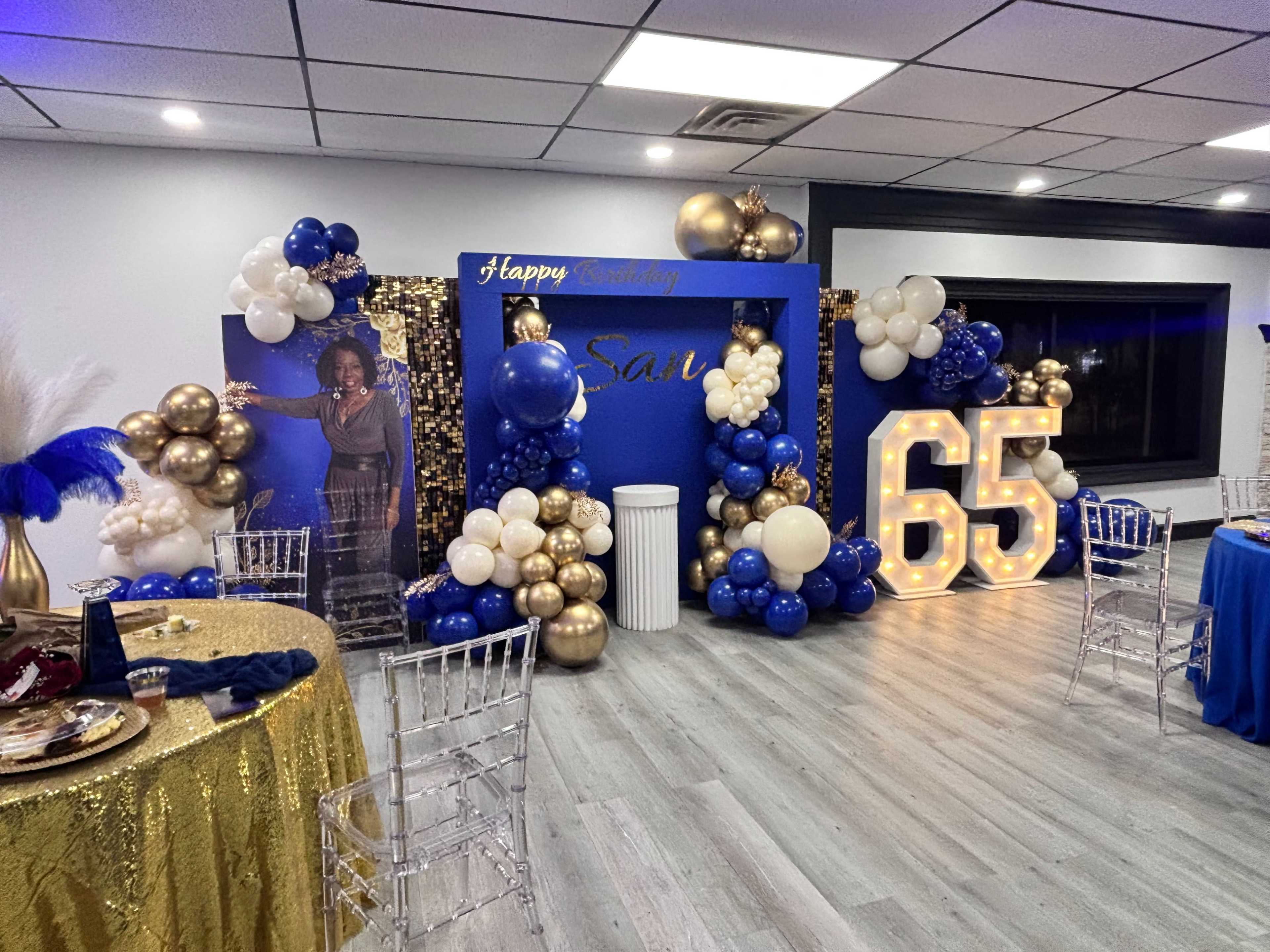The image shows a decorated indoor birthday party scene with a large blue backdrop, golden and white balloons, and a prominent display featuring the number 65.