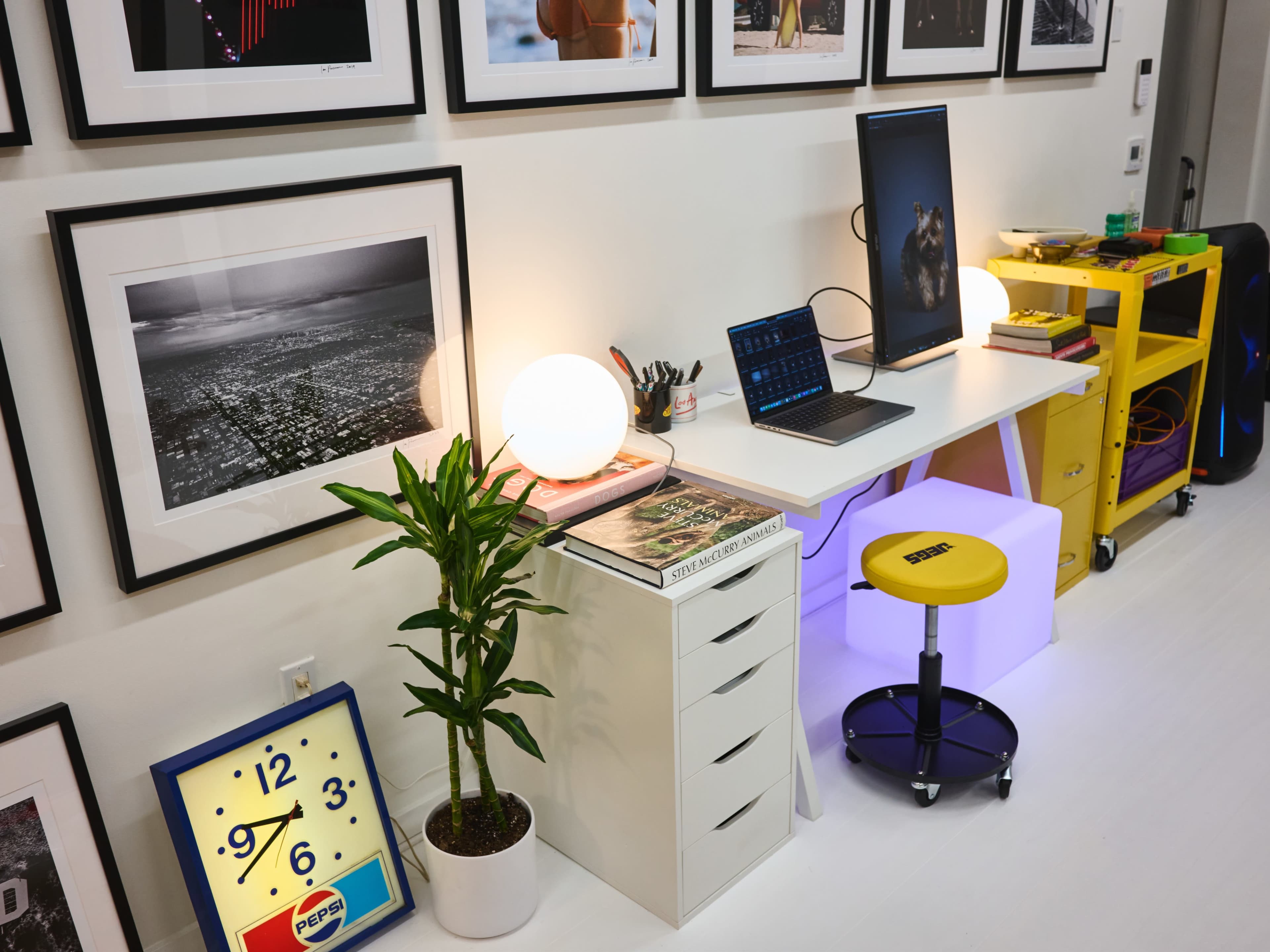 A modern workspace featuring a desk with a computer, decorative plants, a clock, and framed photographs on the wall.